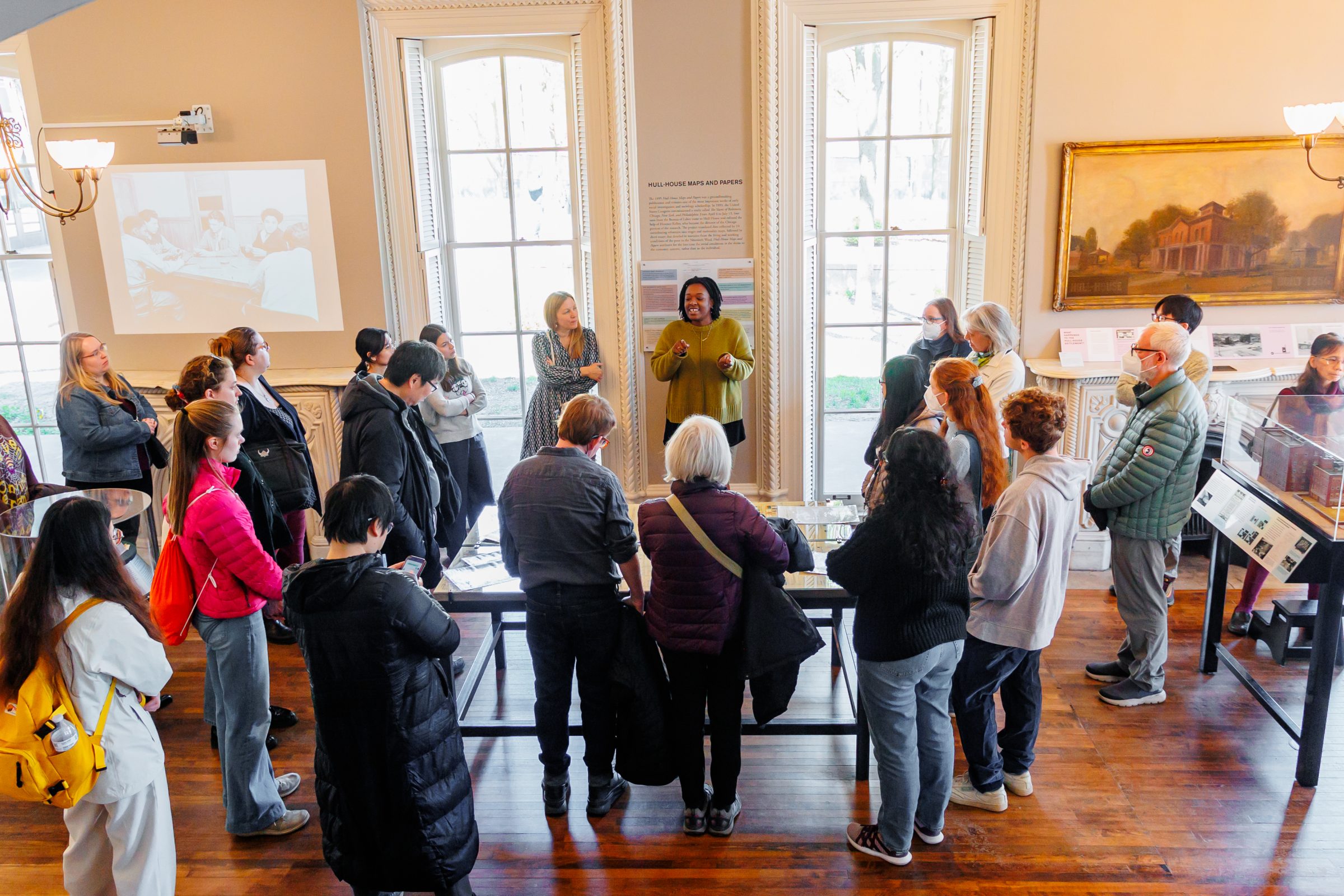 A group of people stands in a museum exhibit room, listening to a speaker in a green outfit. Large windows and historical displays surround the group as they face the speaker.