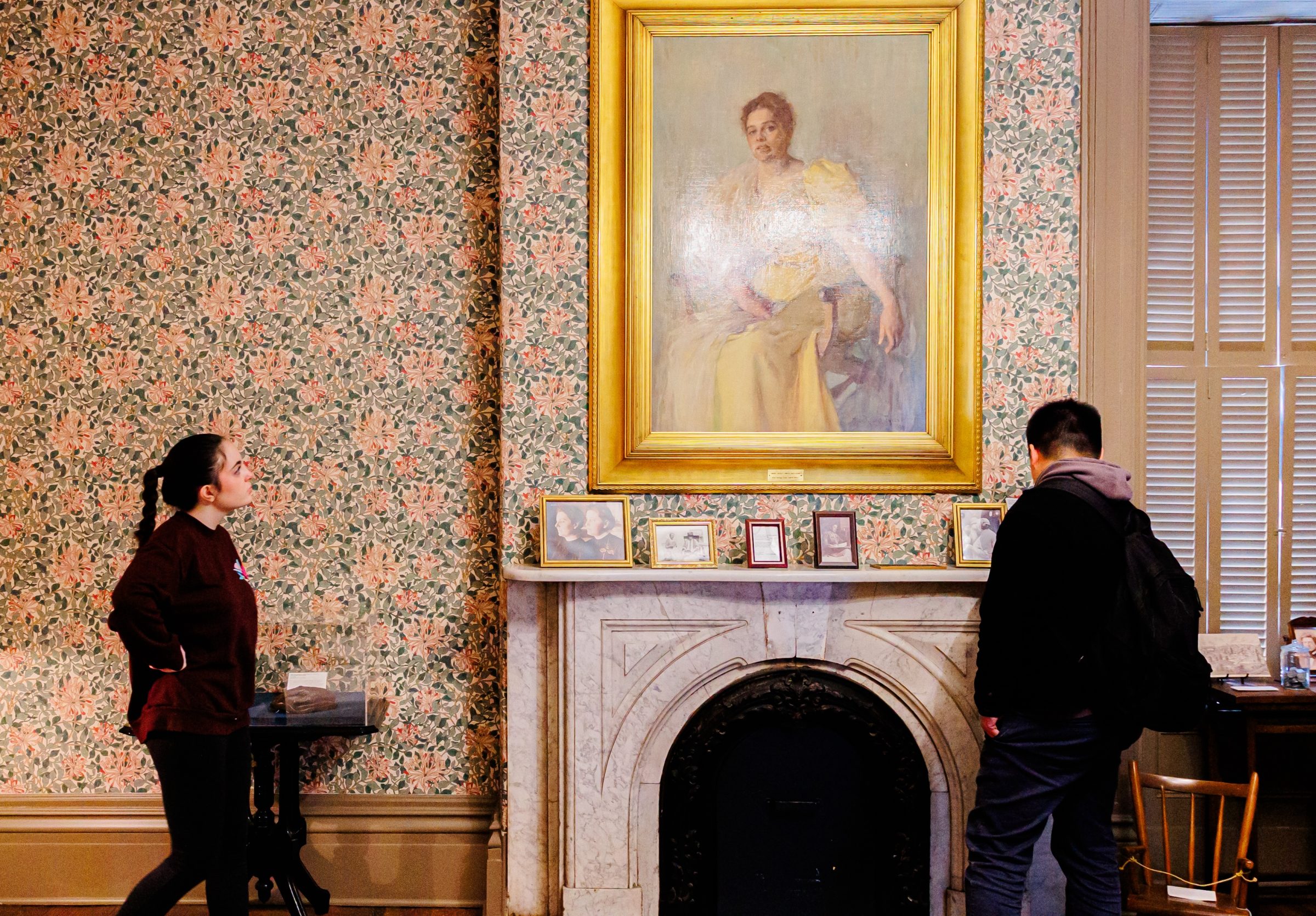 Two museum visitors stand before a marble fireplace in a room with floral wallpaper. Above the mantel hangs a large, ornate portrait of a woman in a yellow dress, with smaller framed photos displayed on the ledge below.