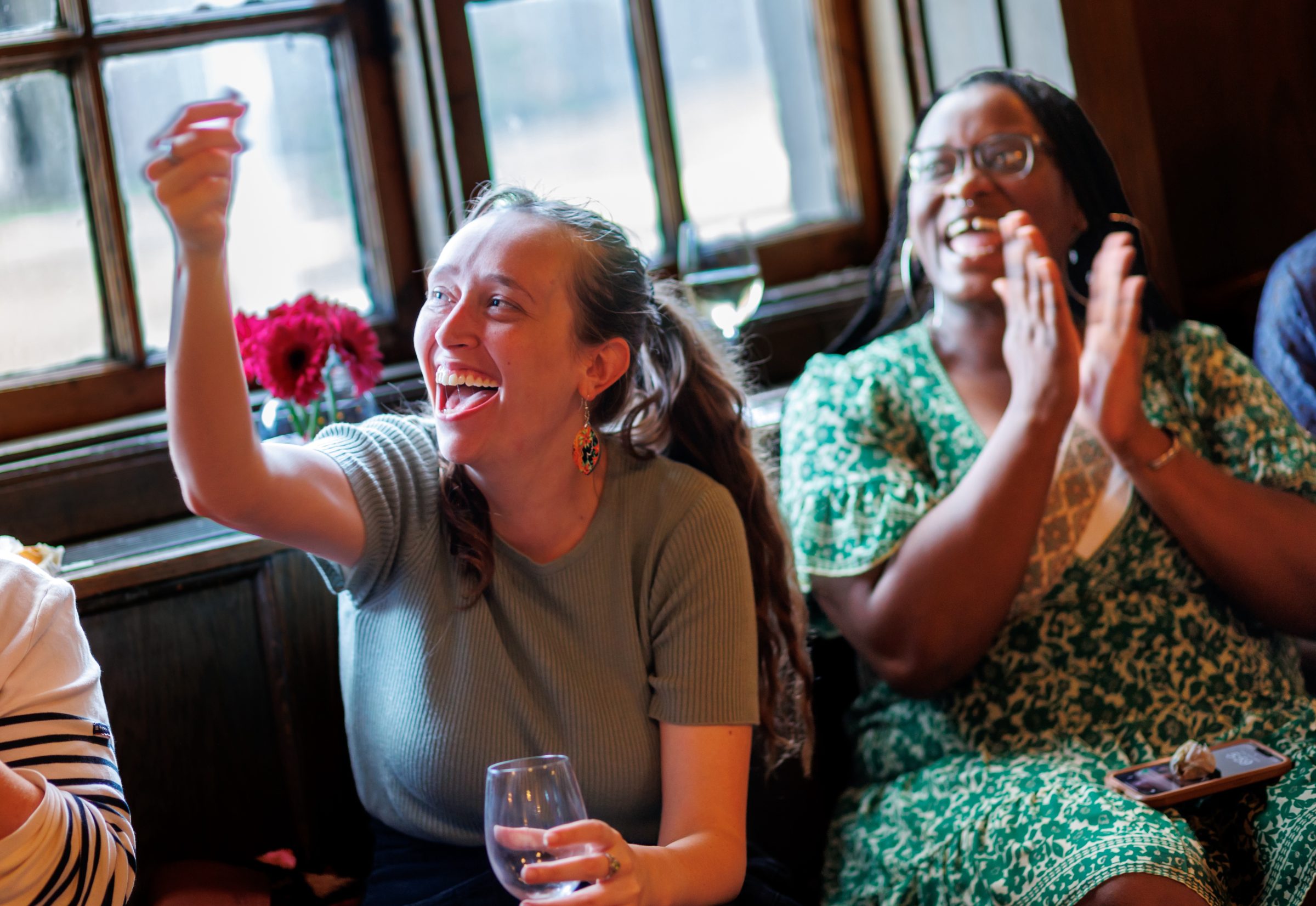 Two women sit indoors by a window, smiling and laughing. One holds a wine glass and gestures excitedly, while the other claps. Both appear to be enjoying a lively social gathering.
