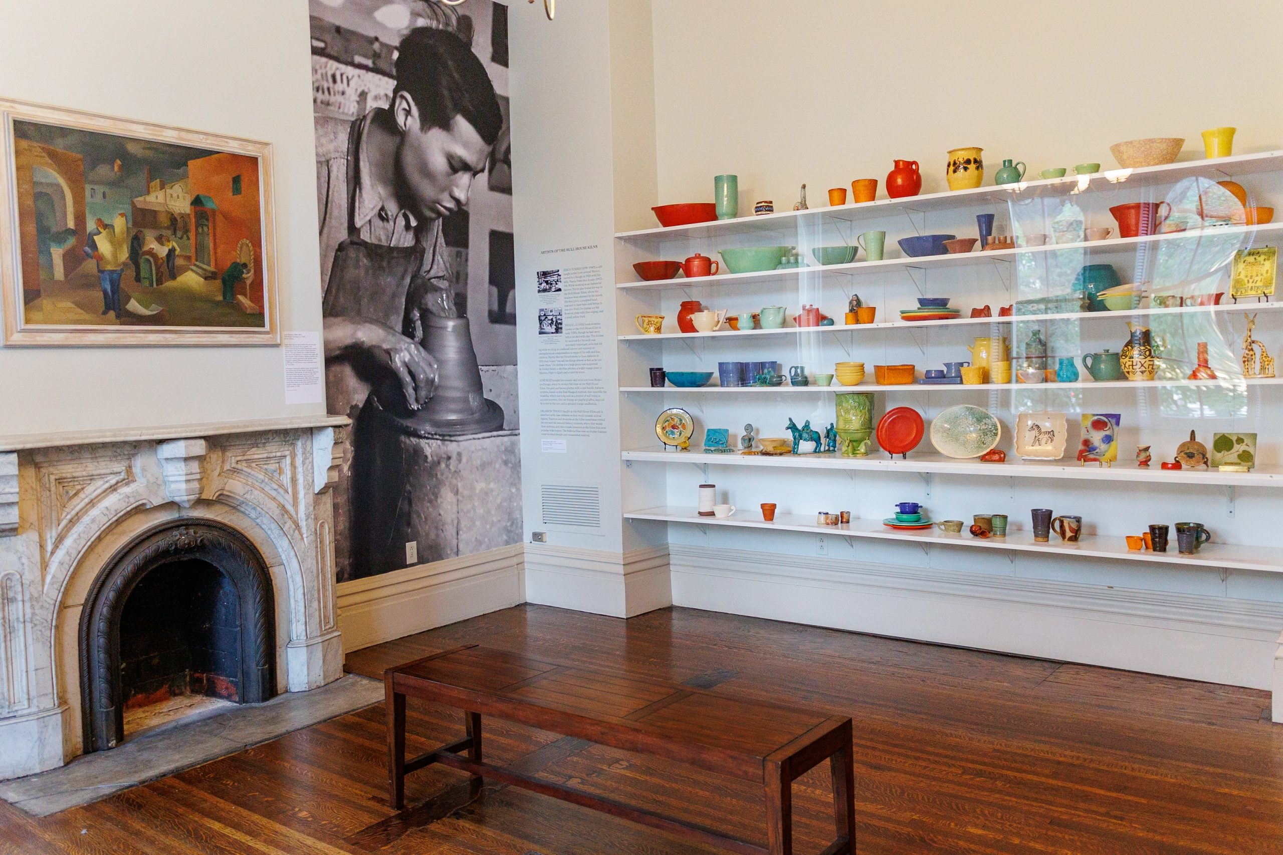 A museum room with wooden floors, a marble fireplace, a wooden bench, colorful pottery displayed on wall shelves, a large black-and-white photo of a potter, and a painting hanging beside the fireplace.