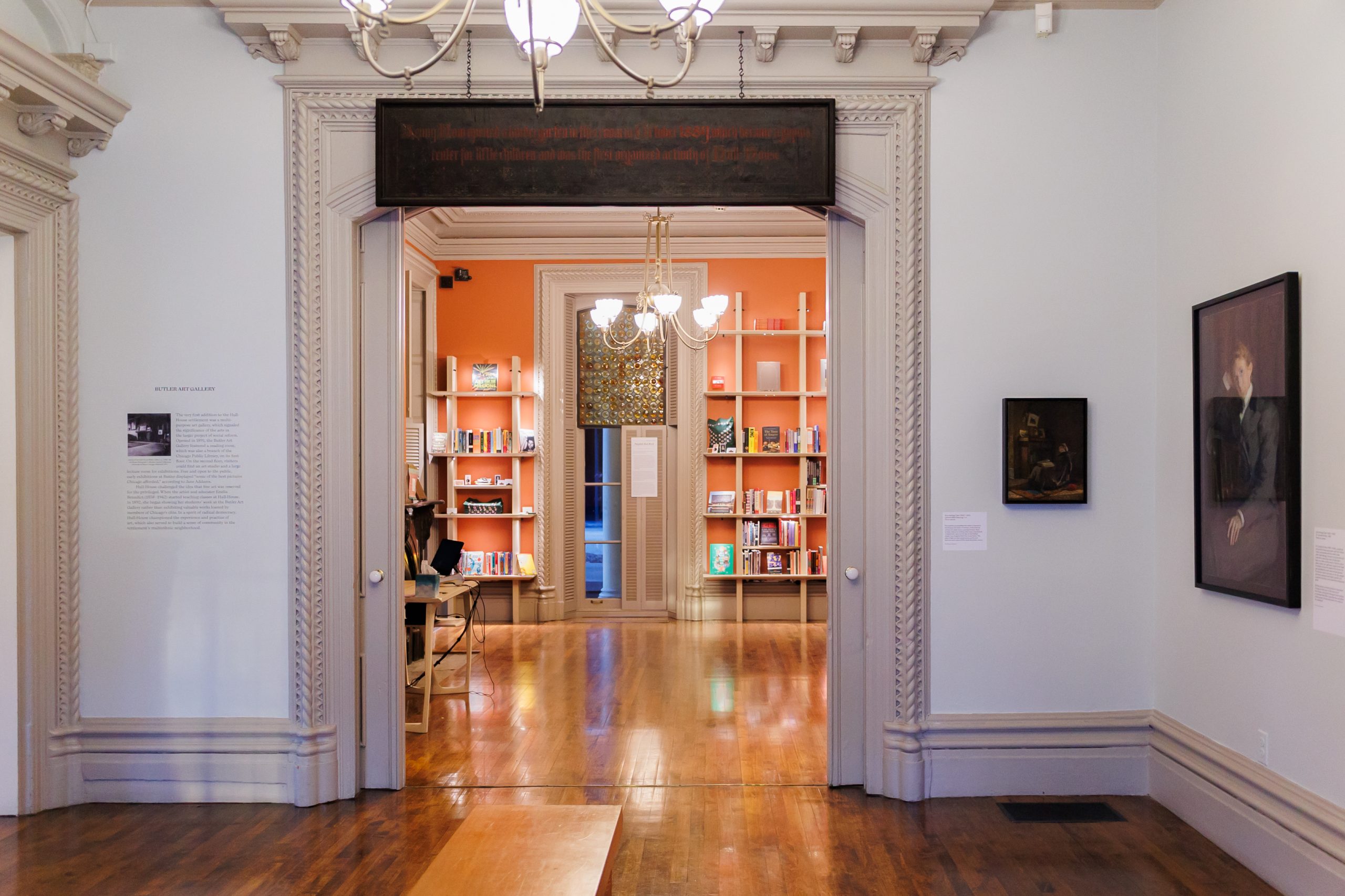 A view through a doorway into a brightly lit room with orange walls and bookshelves; the foreground room has white walls, ornate trim, wood floors, and framed art on the walls.