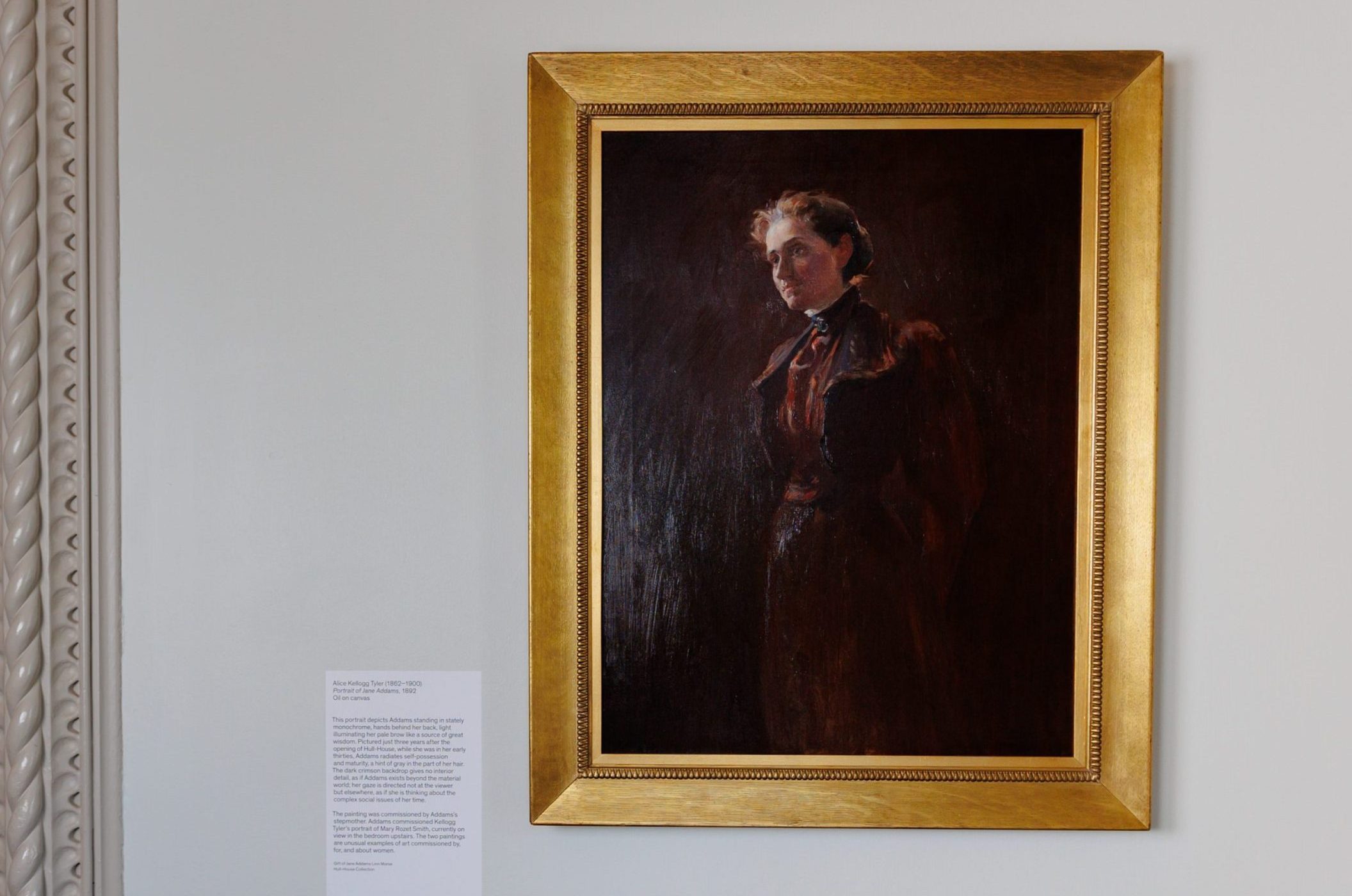 A framed portrait painting of a woman with a serious expression, dressed in dark clothing, hangs on a light-colored wall. An informational plaque with text is mounted next to the artwork.