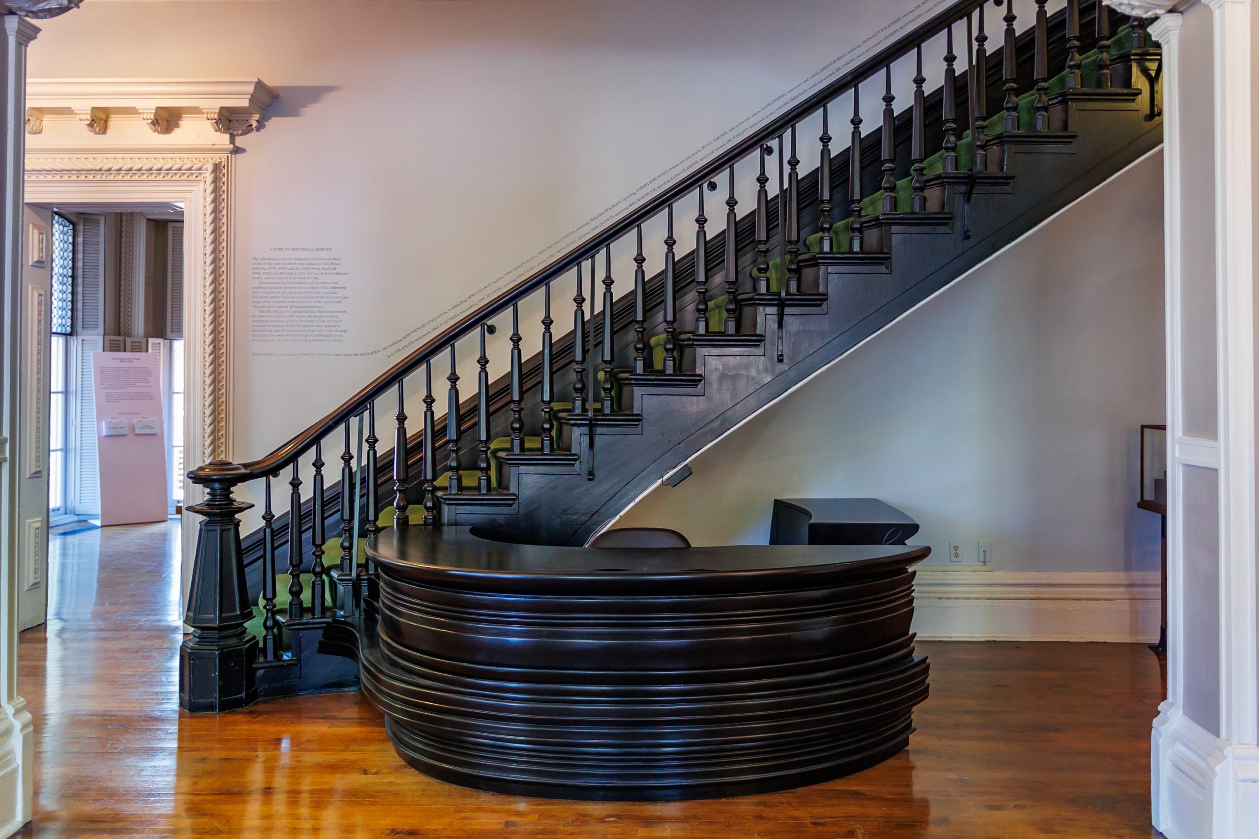 A curved black wooden reception desk sits beneath a staircase with dark railings and green spindles in a room with polished wood floors and ornate trim.