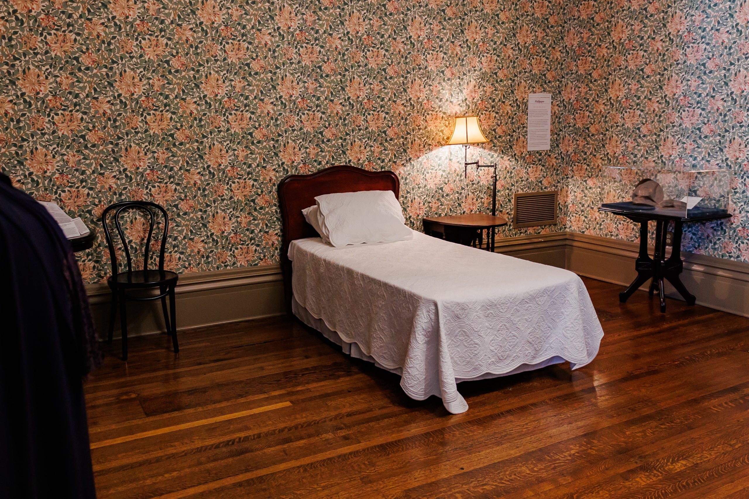 A neatly made single bed with a white quilt is set against a floral wallpapered wall in a vintage-style room. A wooden chair, side table with lamp, and hardwood floors complete the cozy setting.