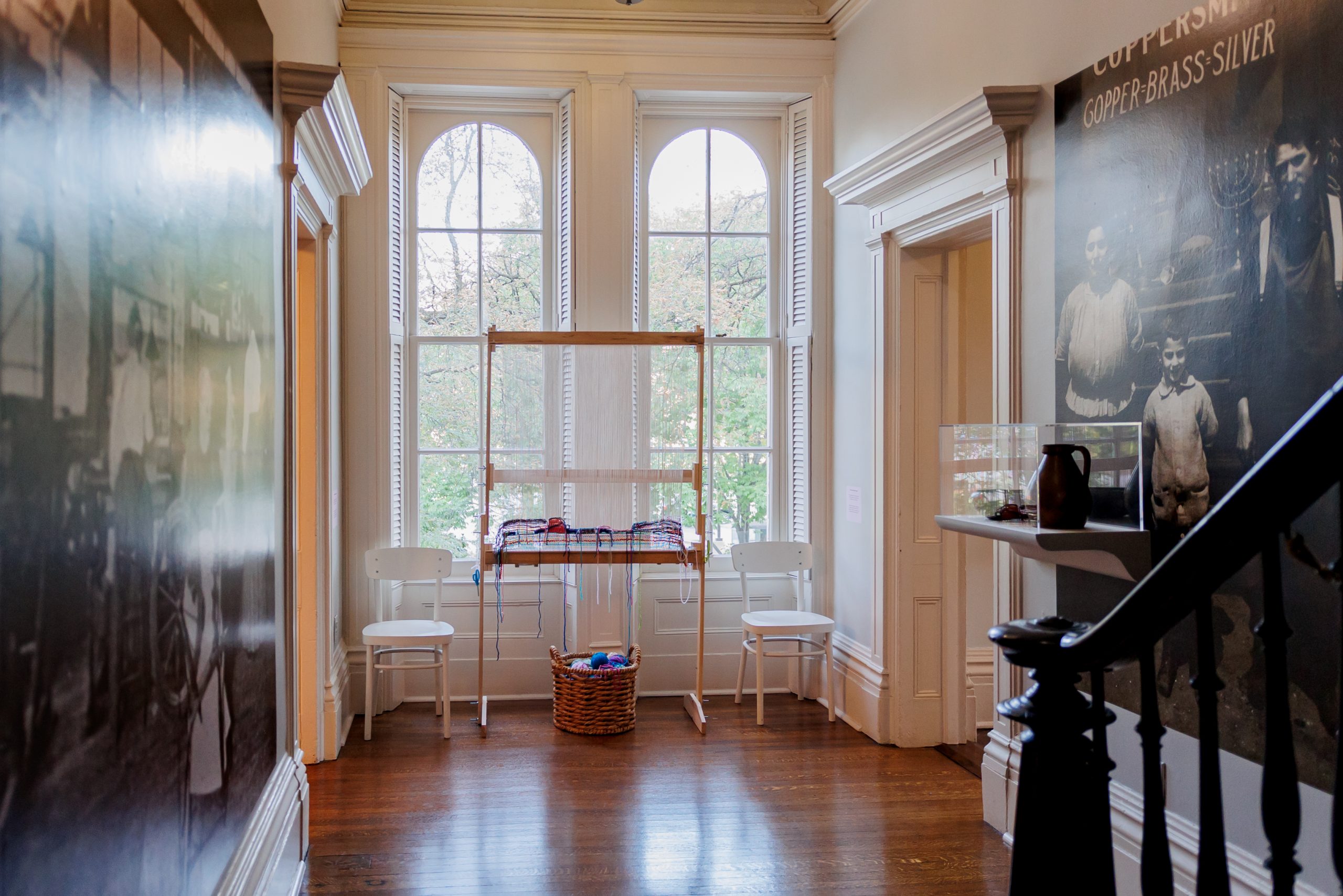 A hallway with large arched windows, two white chairs, a basket, and a loom. Dark wall art and a display case are visible, with wooden floors and staircase banisters in the foreground.
