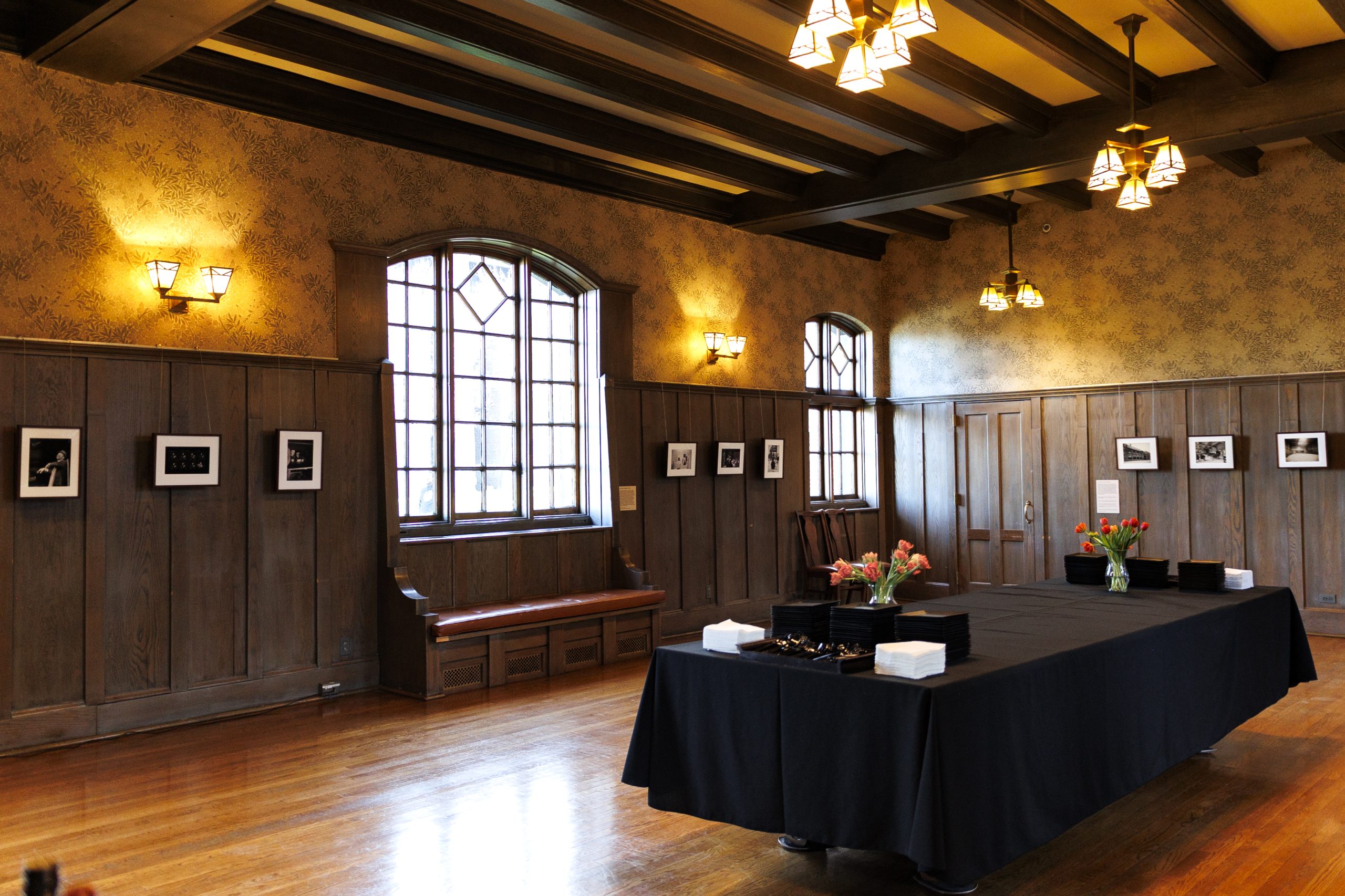 A spacious room with wooden floors, paneled walls, and large windows. Black-and-white photos hang on the walls. Long tables covered in black cloth hold vases of flowers and boxes, beneath hanging lights.