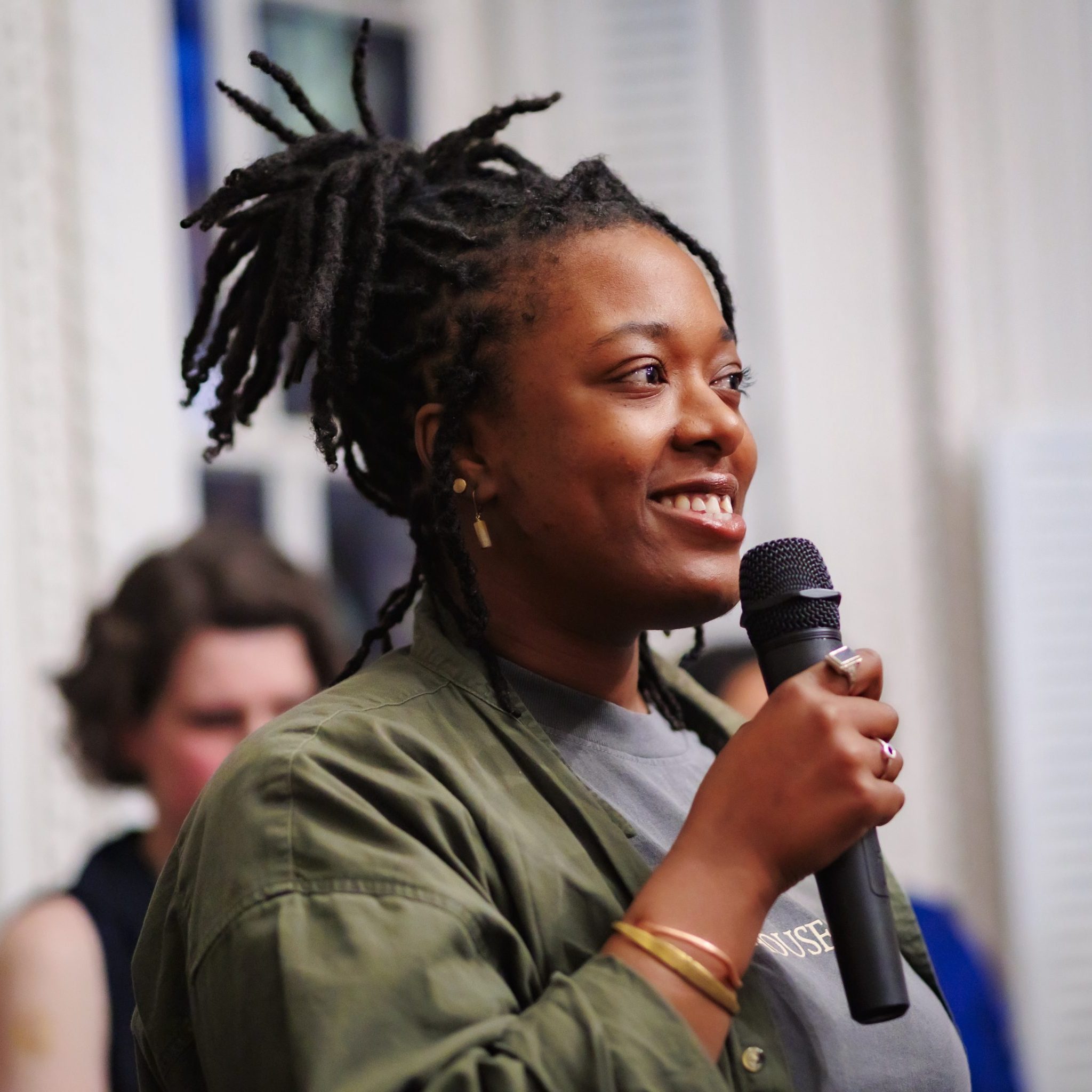 A woman with dark braided hair holds a microphone and smiles while speaking. She wears a green jacket over a gray shirt, with gold earrings and bracelets. The background is blurred with a person and white window shutters visible.