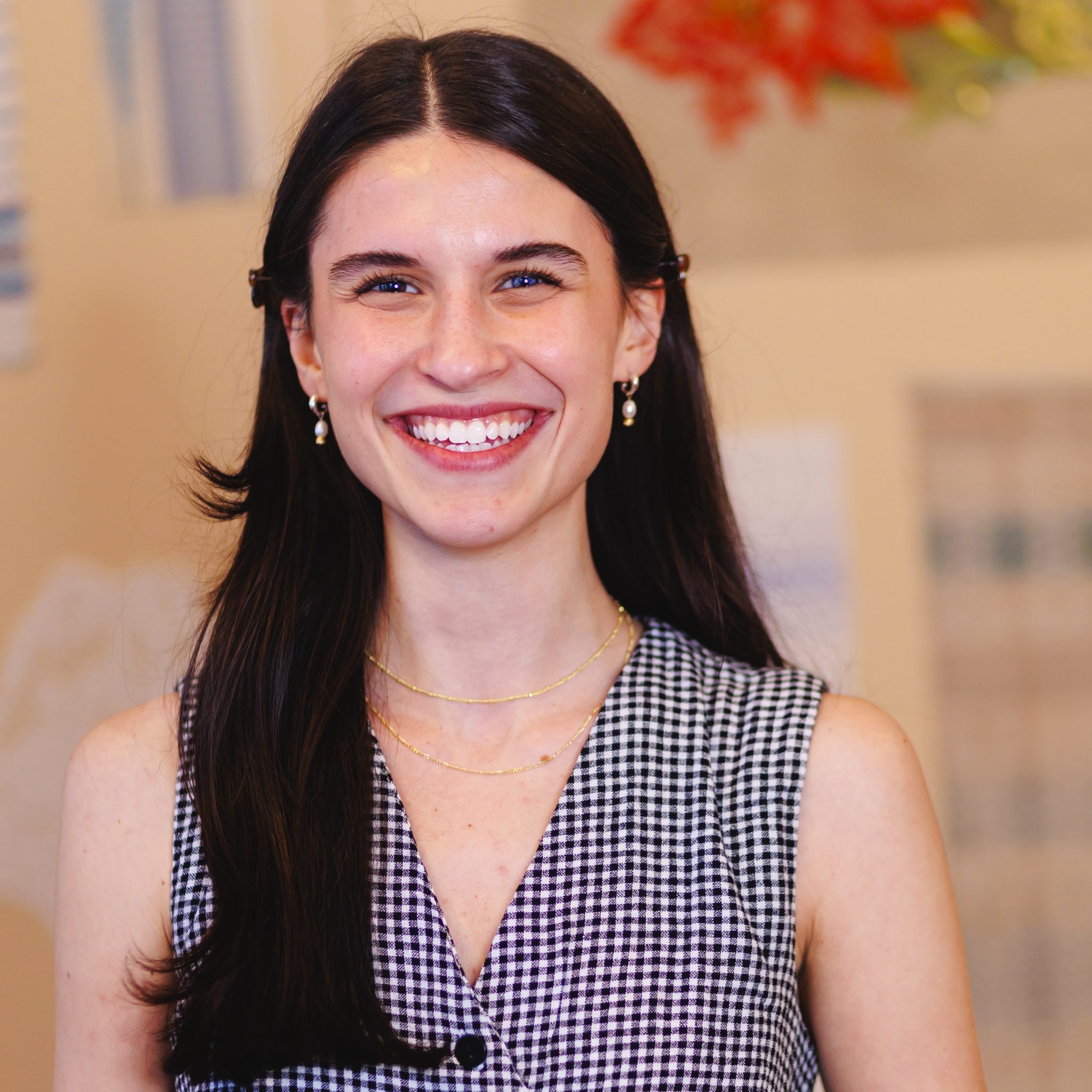 A woman with long dark hair, wearing a sleeveless black-and-white checkered dress and gold jewelry, smiles brightly at the camera. The background is softly blurred with warm colors.