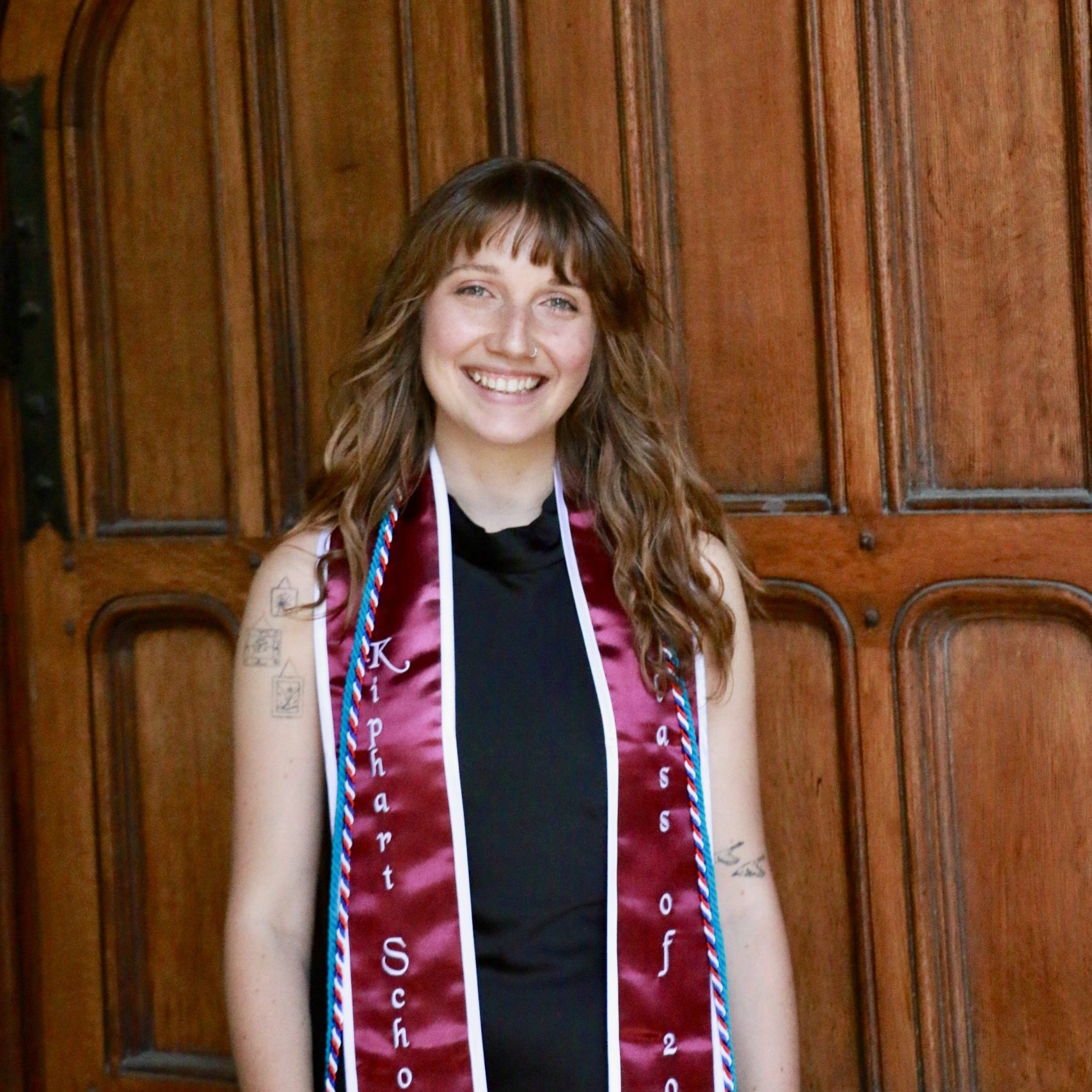 A person with wavy hair in a black outfit and maroon graduation stole smiles in front of a large wooden door.