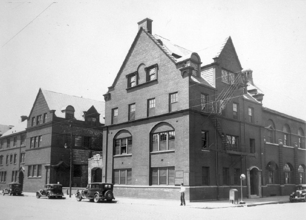 A black-and-white photo shows two large brick buildings with steeply pitched roofs and arched windows. Vintage cars are parked along the street, and a person stands on the sidewalk near a fire escape.