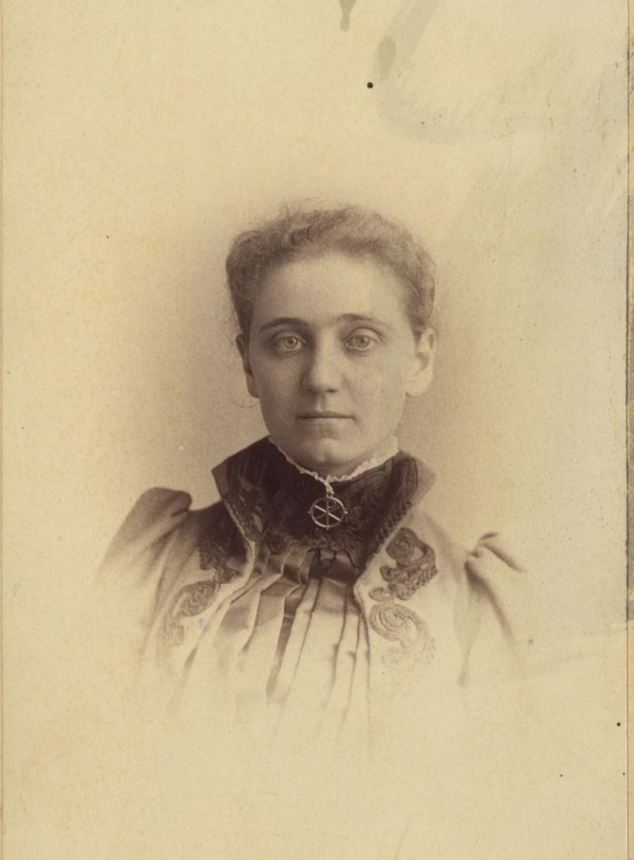 A sepia-toned vintage portrait of a woman with short curly hair, wearing a high-neck dress with embroidered details. The photo is faded around the edges and mounted on a card labeled Max Platz, Chicago.