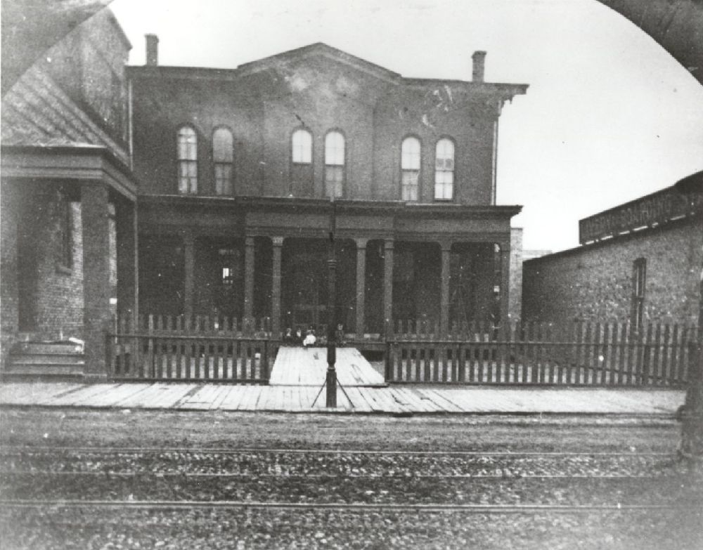 A historic black-and-white photo of a two-story brick building with arched windows, a columned porch, and a picket fence, viewed from across railroad tracks.