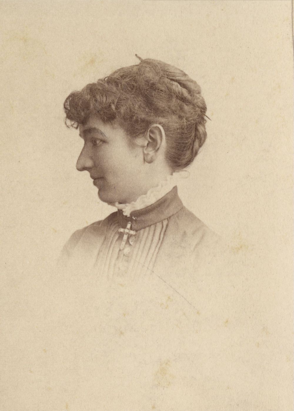 A sepia-toned portrait of a woman in profile, facing left. She has curly hair pinned up, is wearing a high-collared blouse with a decorative brooch, and has a calm, composed expression. The background is plain and faded.