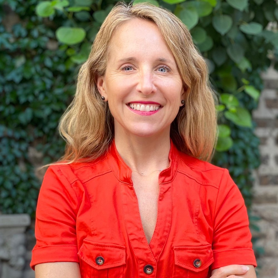 A smiling woman with long blonde hair wearing a bright red blouse stands outdoors with her arms crossed, in front of a background of green leafy plants and a brick wall.
