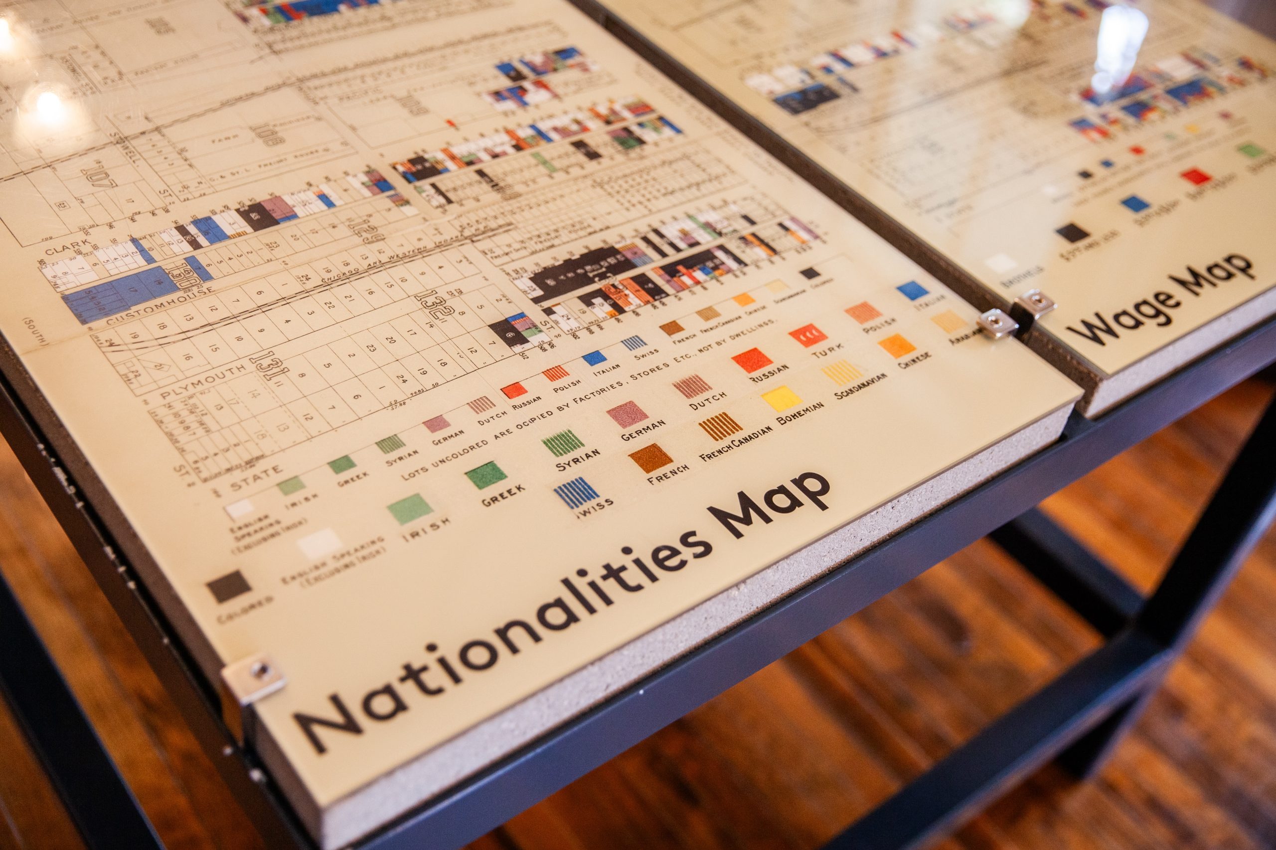 A display table shows a Nationalities Map with colorful boxes representing different countries, and part of a Wage Map beside it. The maps use charts and legends to present information. The table stands on a wooden floor.