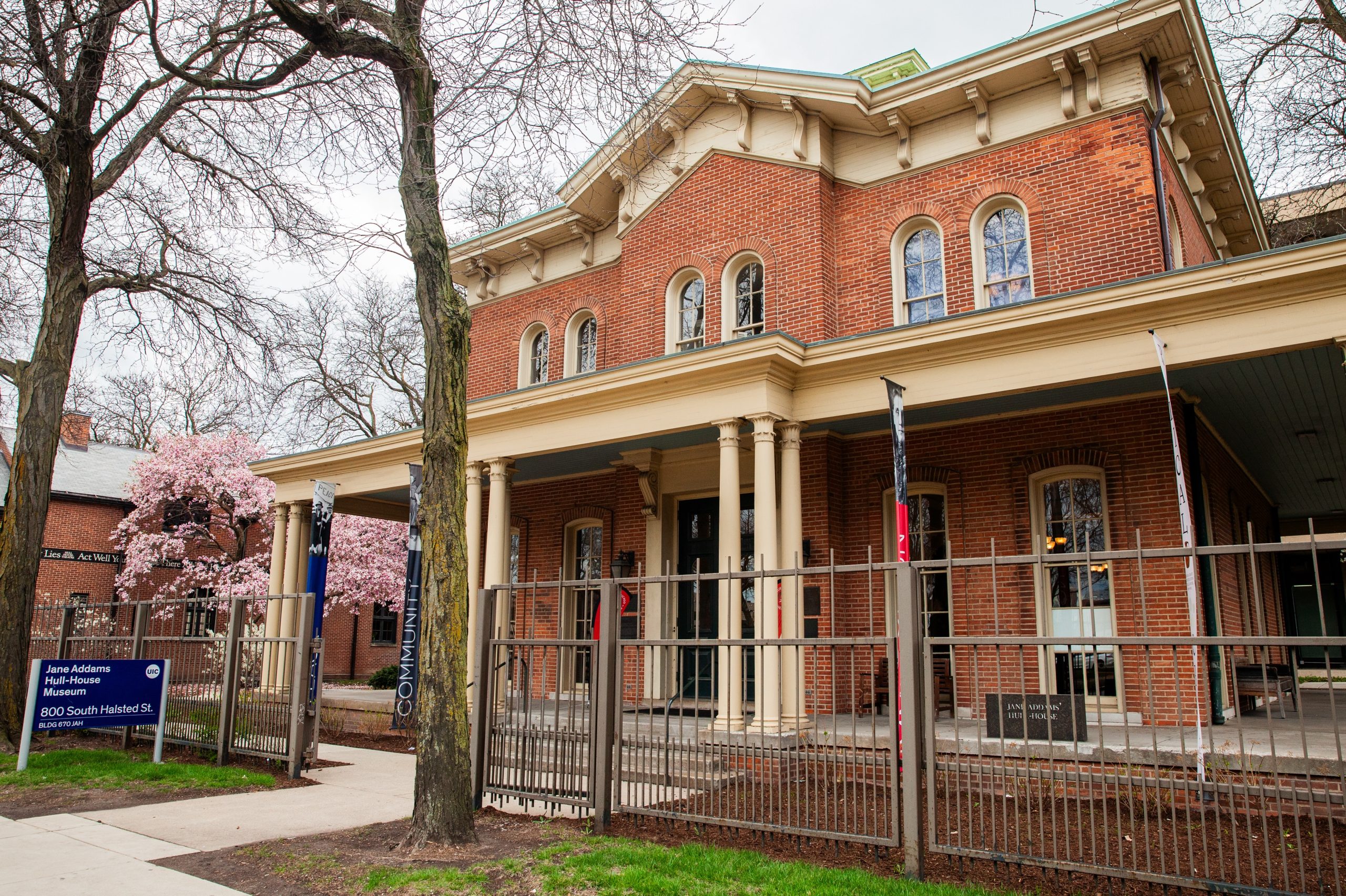 A historic brick building with tall columns, arched windows, and a fenced front yard. A sign reads Jane Addams Hull-House Museum, 800 South Halsted. Trees with spring blossoms are visible nearby.