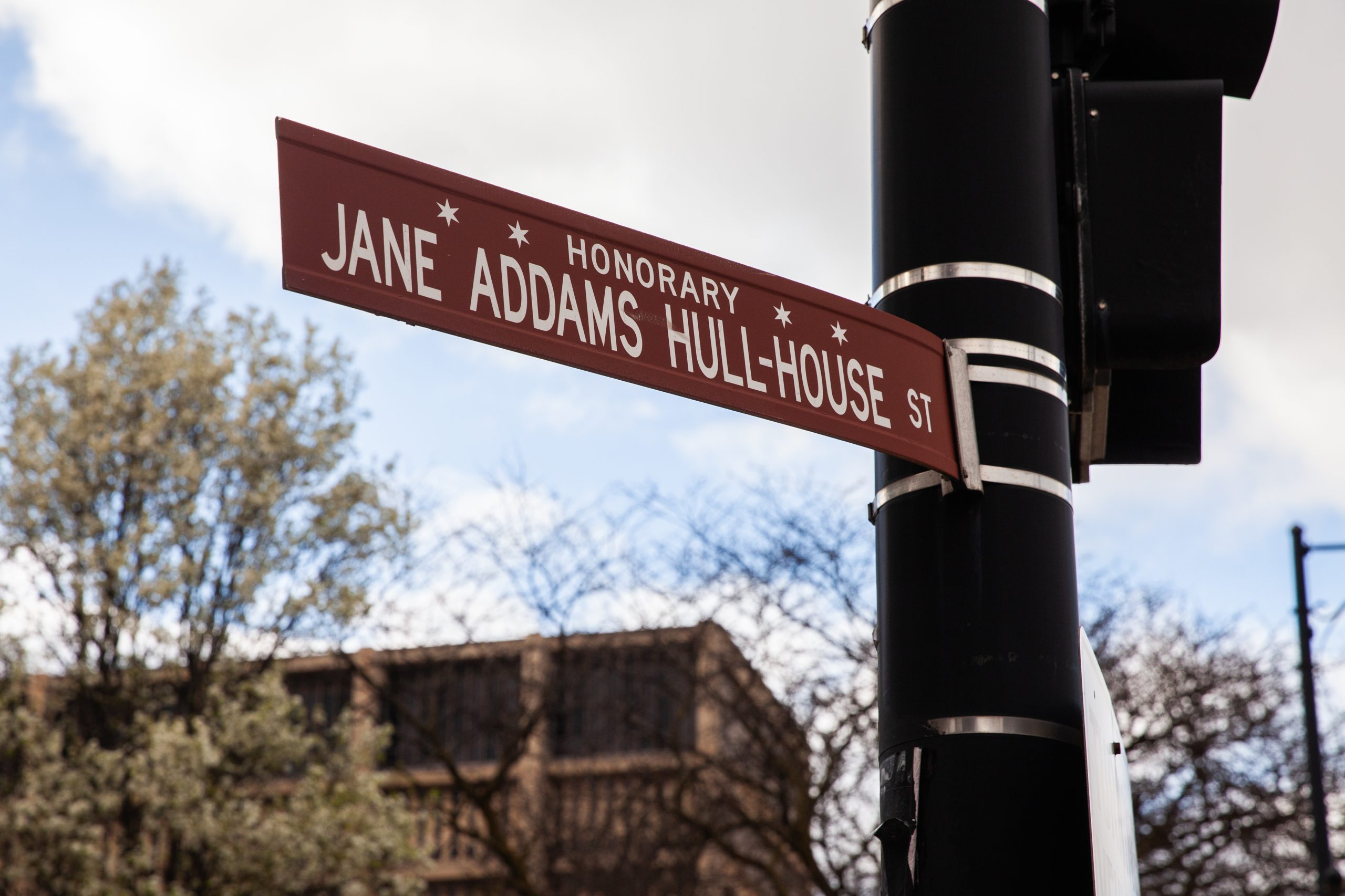 A red street sign reading Honorary Jane Addams Hull-House St is mounted on a black pole, with trees and a building visible in the background.
