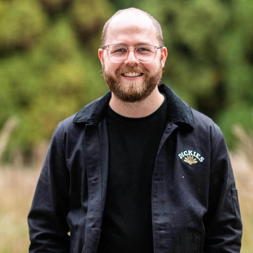 A bearded man with glasses smiles, standing outdoors in front of blurred greenery. He wears a black Dickies jacket over a black shirt.