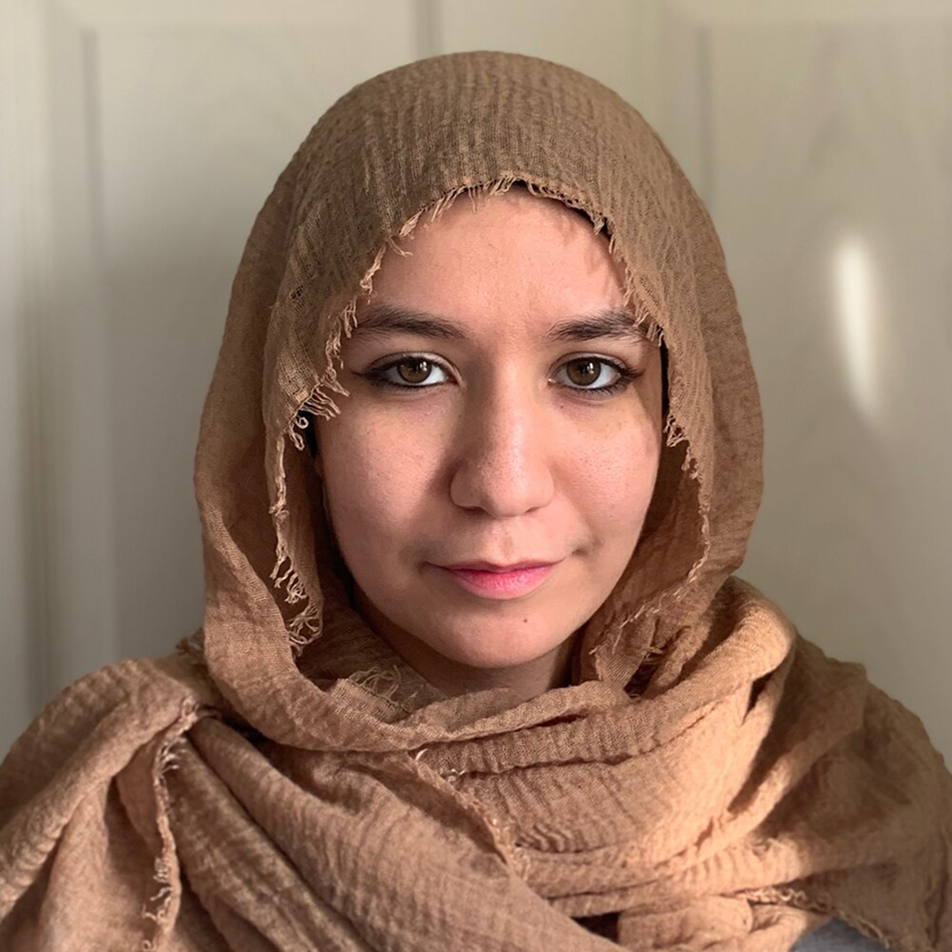 A woman wearing a light brown textured hijab looks directly at the camera with a neutral expression, standing in front of a softly lit, light-colored background.