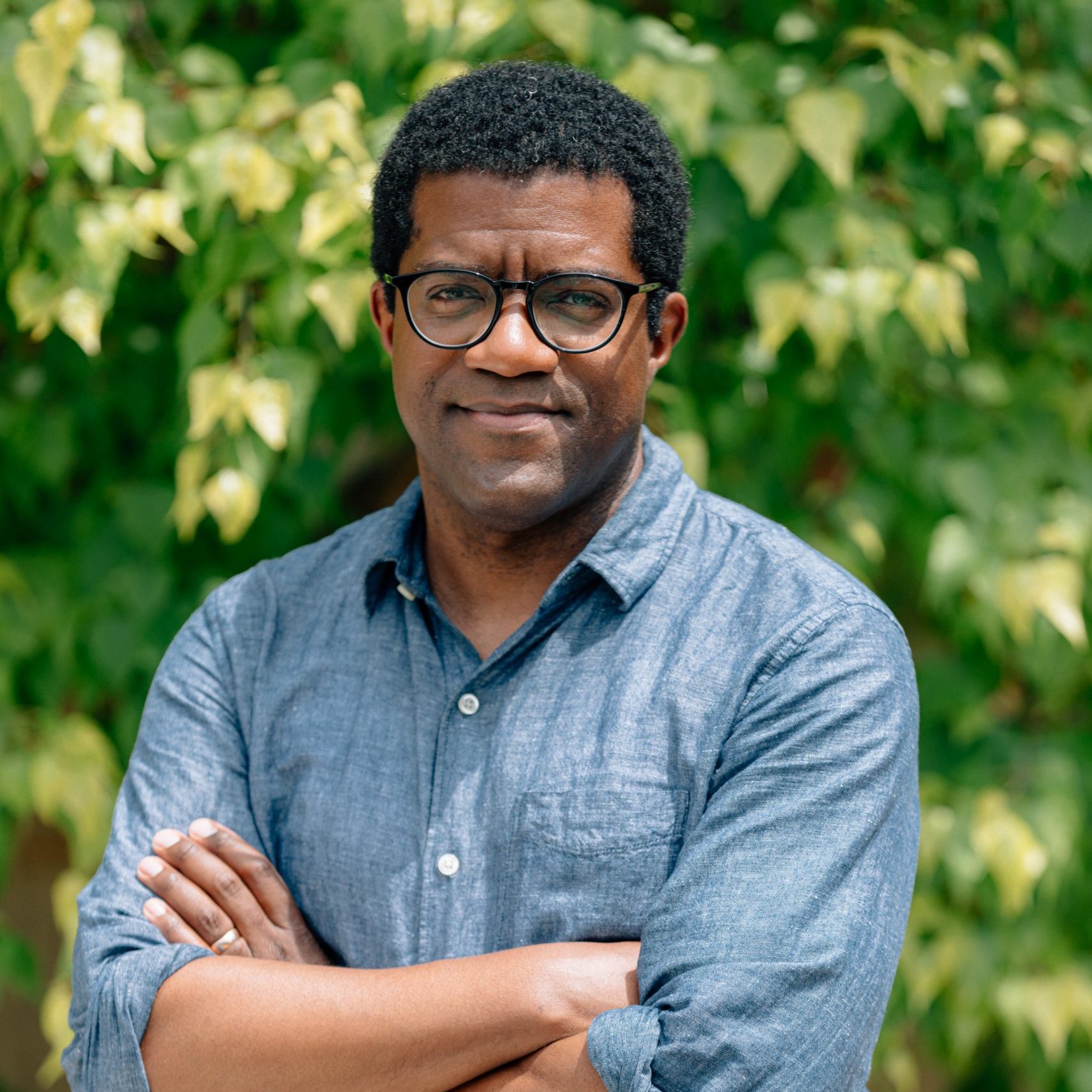 A man wearing glasses and a blue button-up shirt stands outdoors with his arms crossed, in front of green leafy trees.