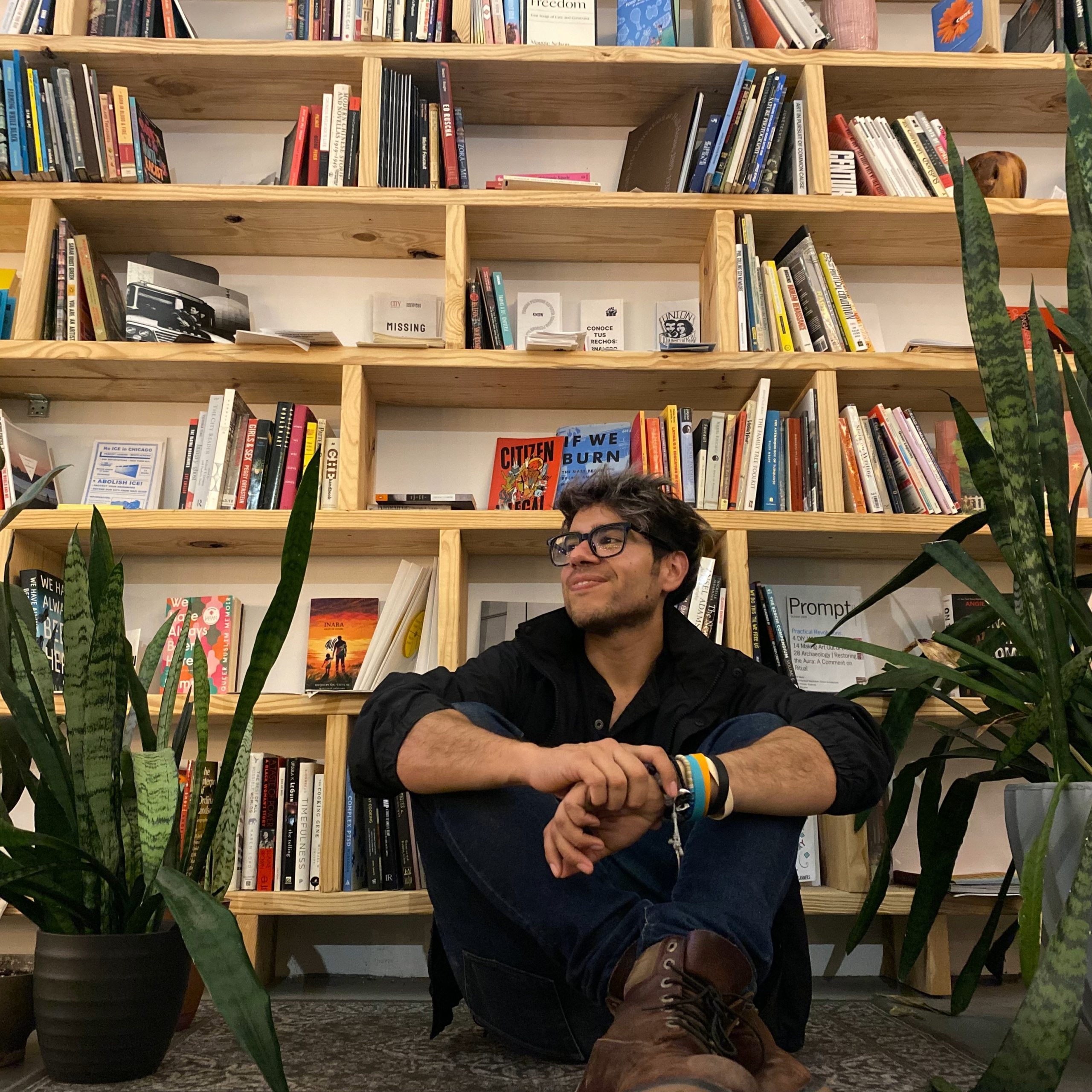 A person wearing glasses and a black shirt sits cross-legged on the floor, smiling, in front of a filled wooden bookshelf, surrounded by green potted plants.