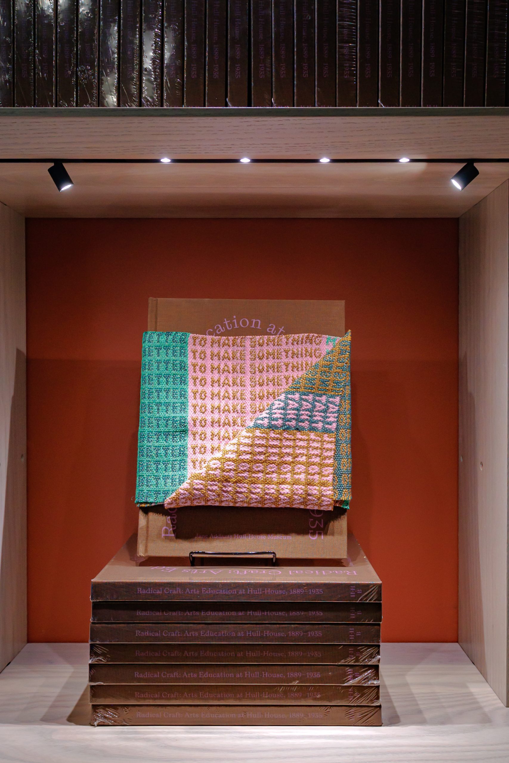 A stack of books sits on a shelf with one open book upright, partially covered by a colorful woven fabric that says TO MAKE SOMETHING BETTER in repeated lines. The shelf is lit by three small spotlights above.