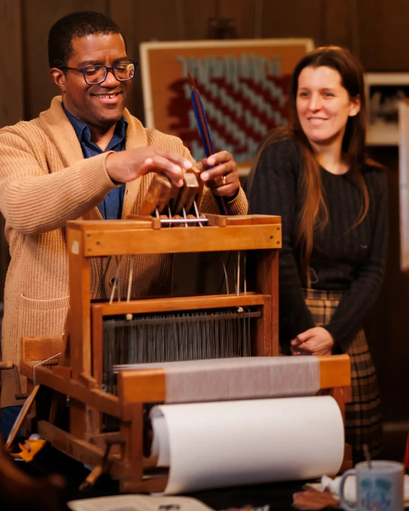A man smiles while using a wooden weaving loom, with a woman standing beside him watching and smiling. The background shows textile art and warm indoor lighting.