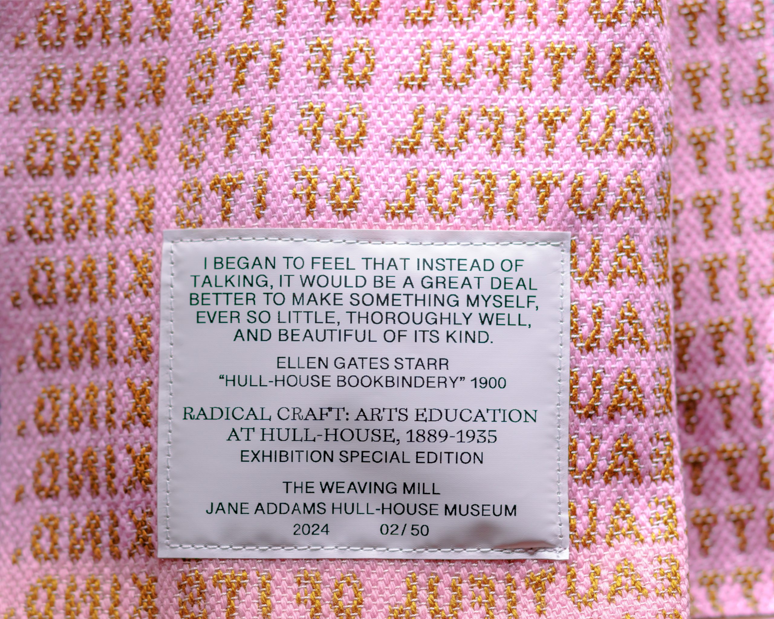 A pink woven fabric with repeating gold text displays a stitched white label. The label features a quote by Ellen Gates Starr, exhibition details for Radical Craft, and credits The Weaving Mill and Hull-House Museum.