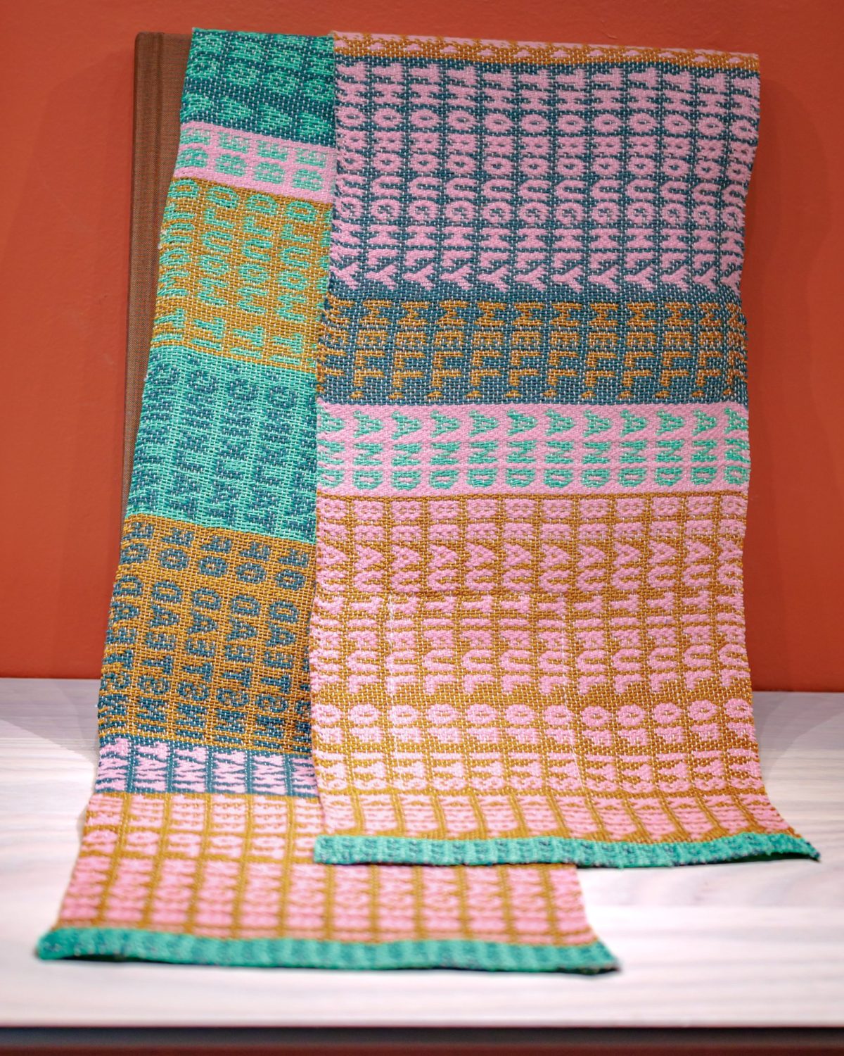 A colorful knitted scarf with geometric patterns in pink, green, mustard yellow, and blue, draped over a wooden stand against an orange background.