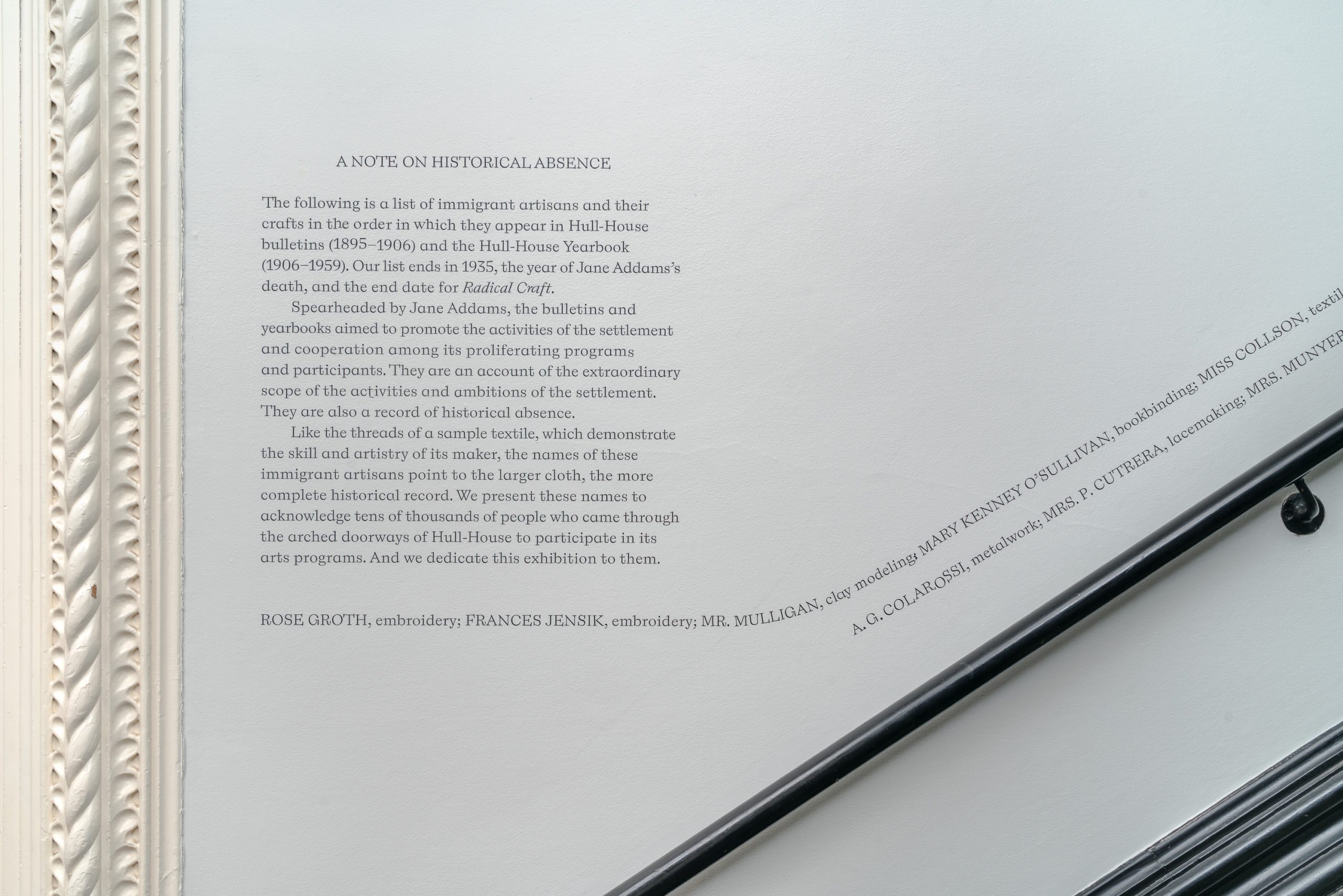 Black text on a white wall explains the historical absence of immigrant artisans from official records, despite their contributions. Ornate white molding frames the left side of the wall.