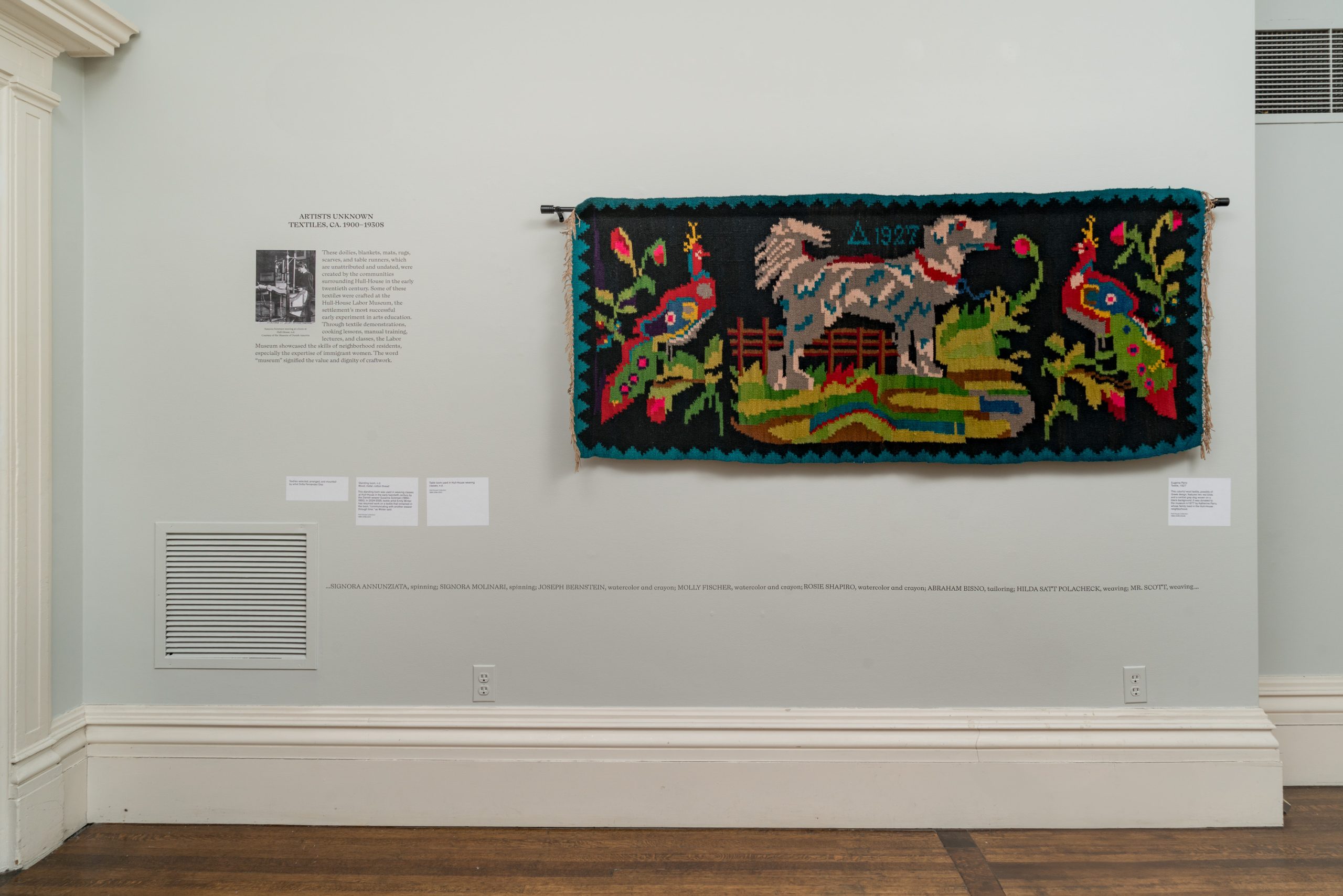 A colorful woven tapestry featuring a stylized animal, plants, and the text “Δ 1927” hangs on a light gray wall in a gallery, accompanied by informational text panels and labels.