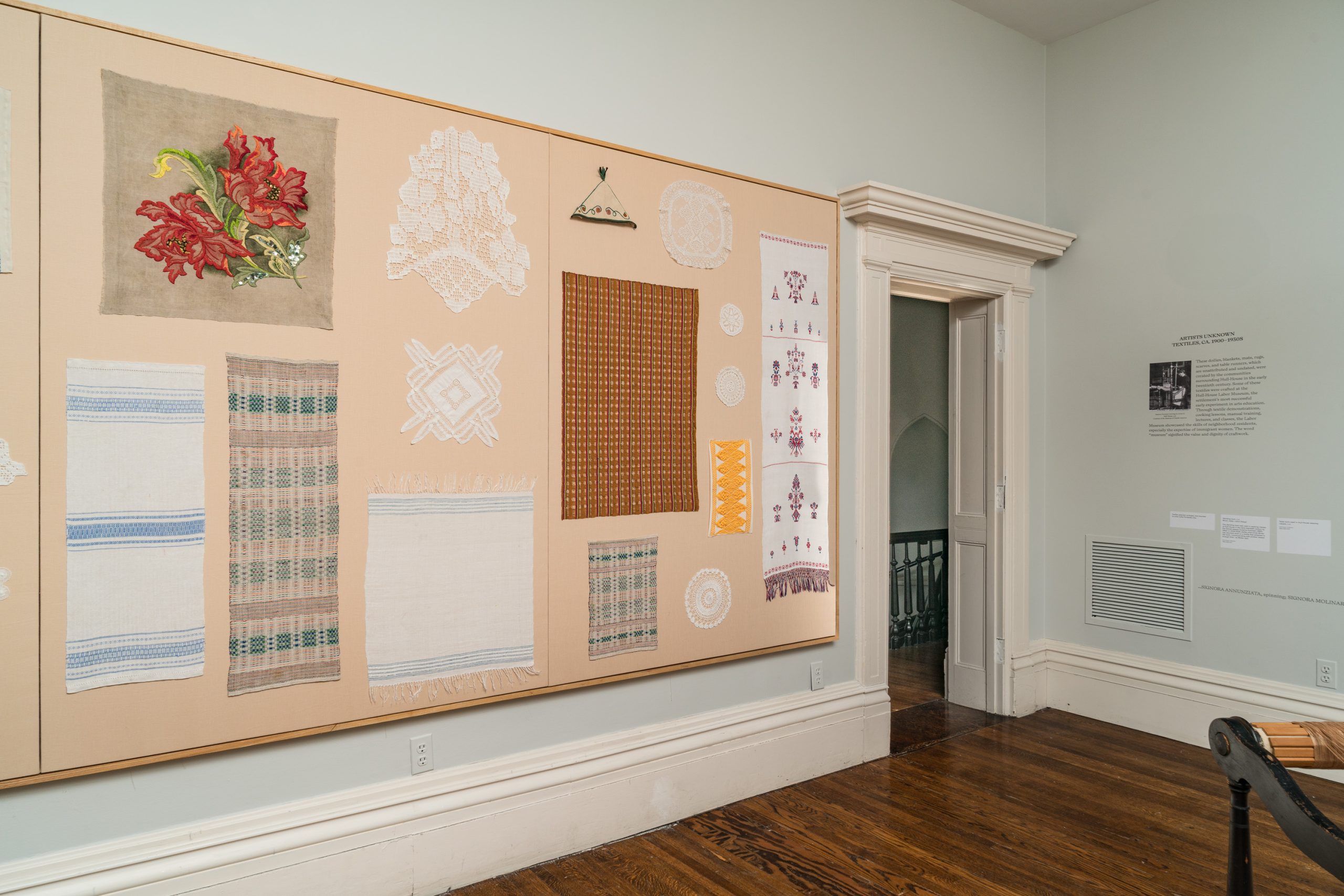 A museum display wall features various textile pieces, including embroidered flowers, woven patterns, lace, and doilies. The room has pale walls, wooden floors, and a doorway leading to another space.