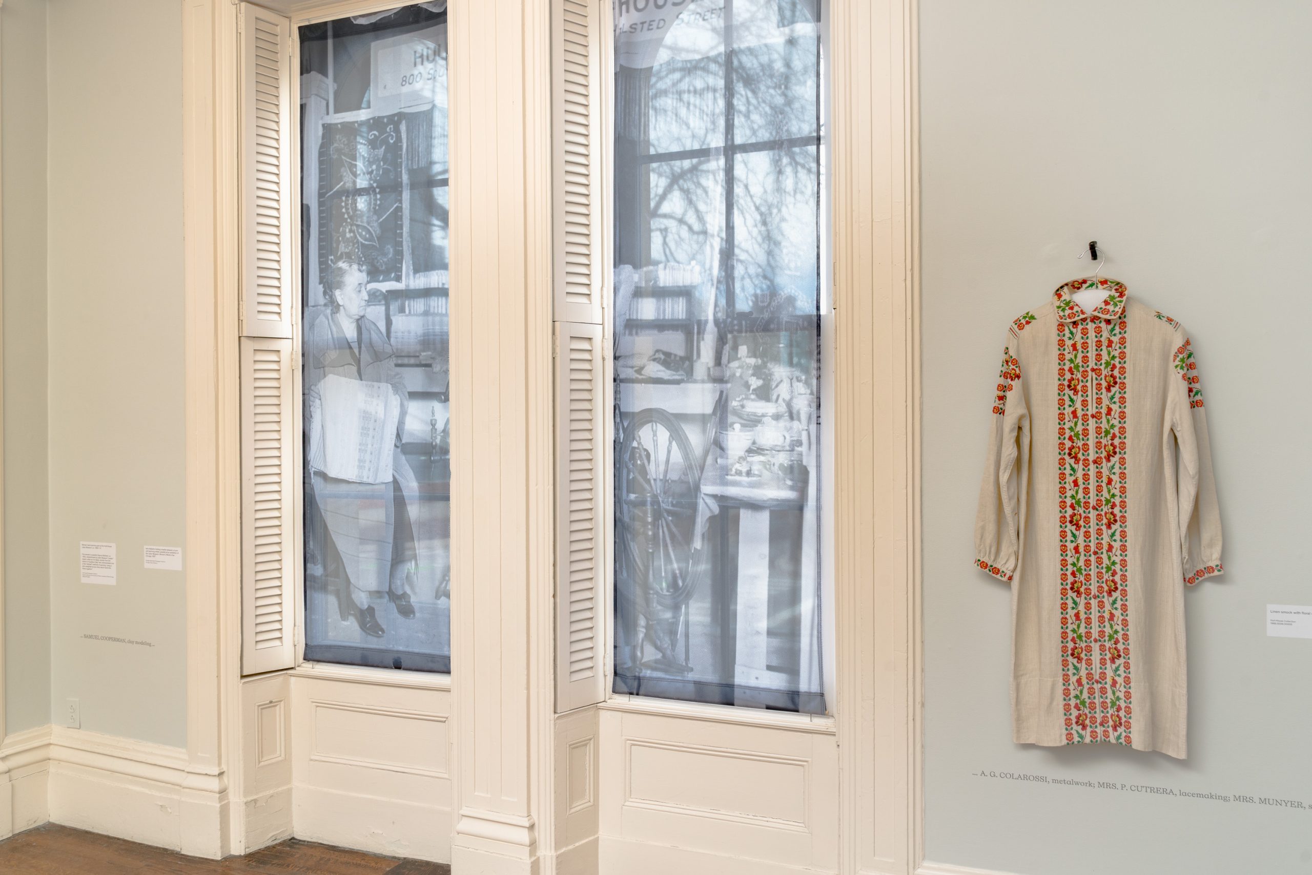 A light-filled room with two tall windows covered in black-and-white photos, showing vintage scenes. To the right, a traditional long-sleeved embroidered garment hangs on the wall.