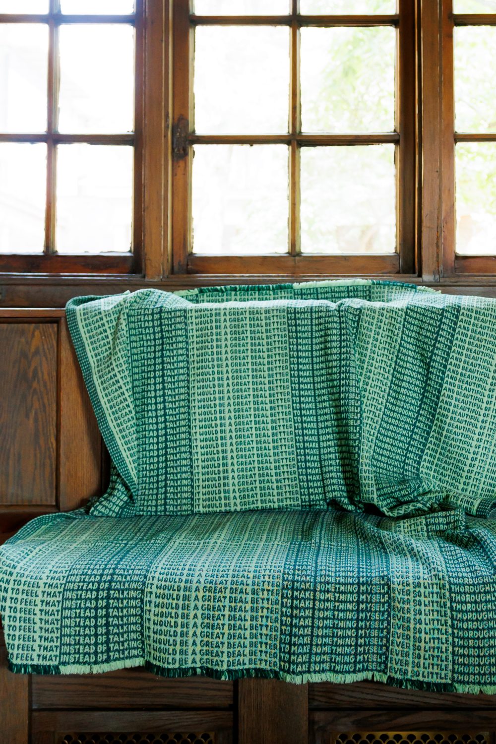 A wooden bench with a green and blue blanket draped over it, displaying repeated text that says, “IT WOULD BE A GREAT DEAL TO TALK THAT TALK.” Sunlight streams through windowpanes in the background.