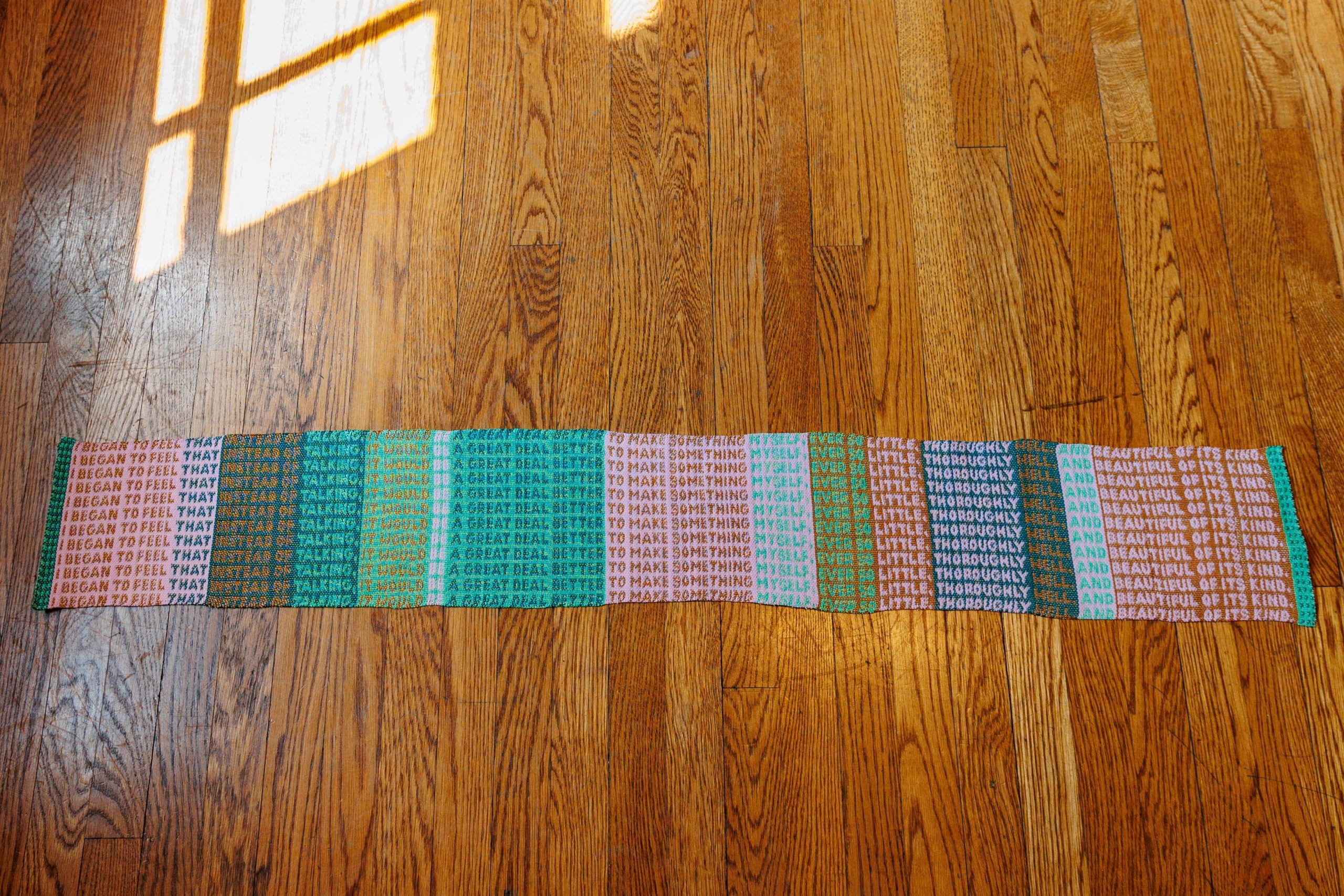 A colorful, rectangular scarf with repeated text patterns lies flat on a wooden floor. Sunlight streams onto the floor from the left side, casting bright reflections and shadows.