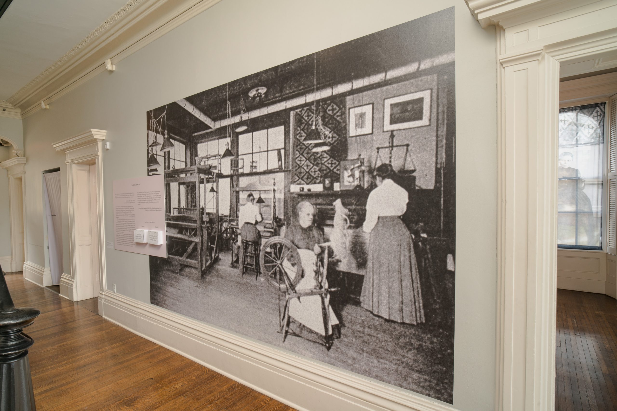 A large black-and-white historical photograph displayed on a museum wall shows women working in a textile workshop, with wooden floors and decorative moldings visible in the surrounding gallery space.