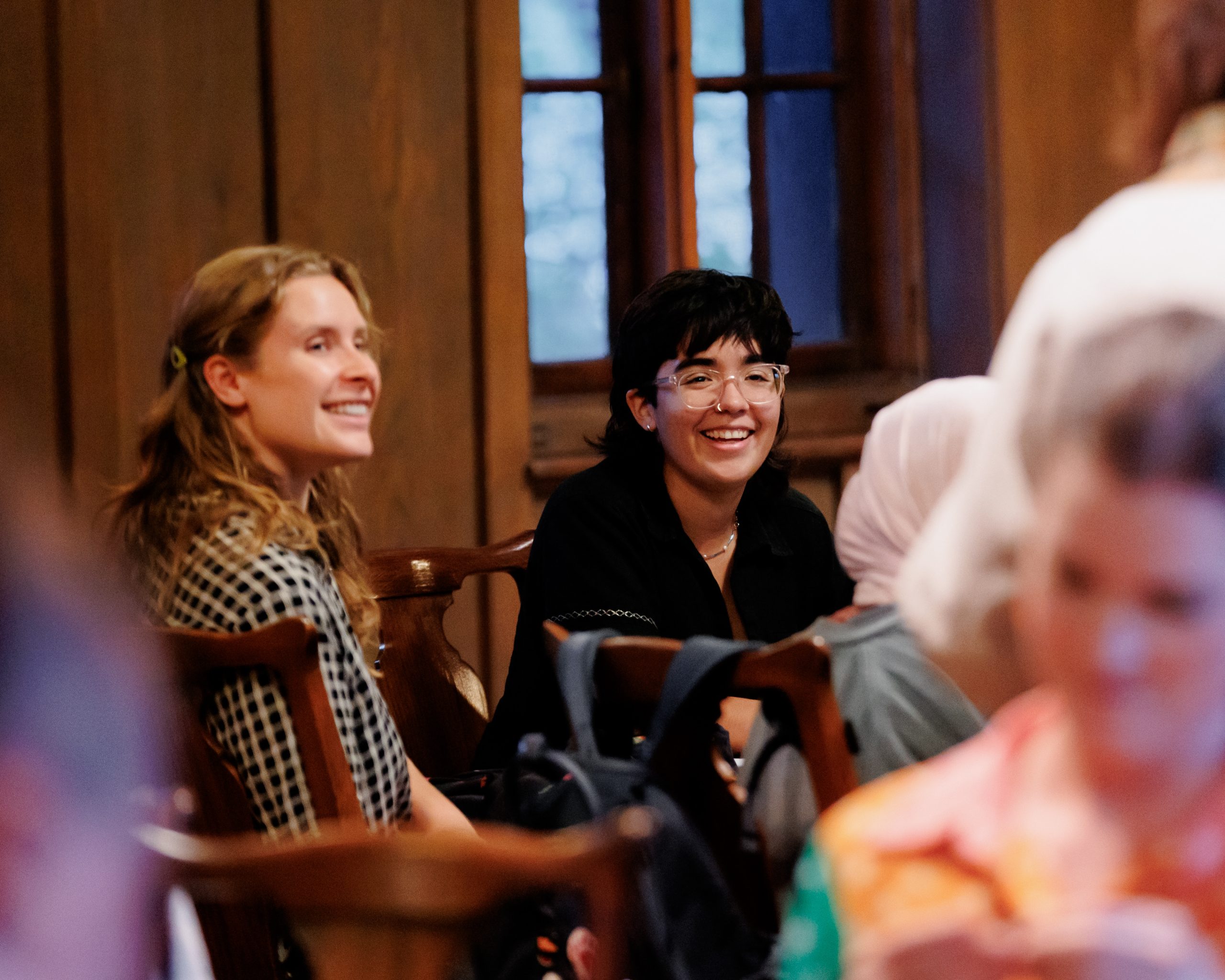 Two people are sitting indoors on wooden chairs, smiling and looking toward the camera. The background features wooden walls and a window. Other people are present, but out of focus in the foreground.