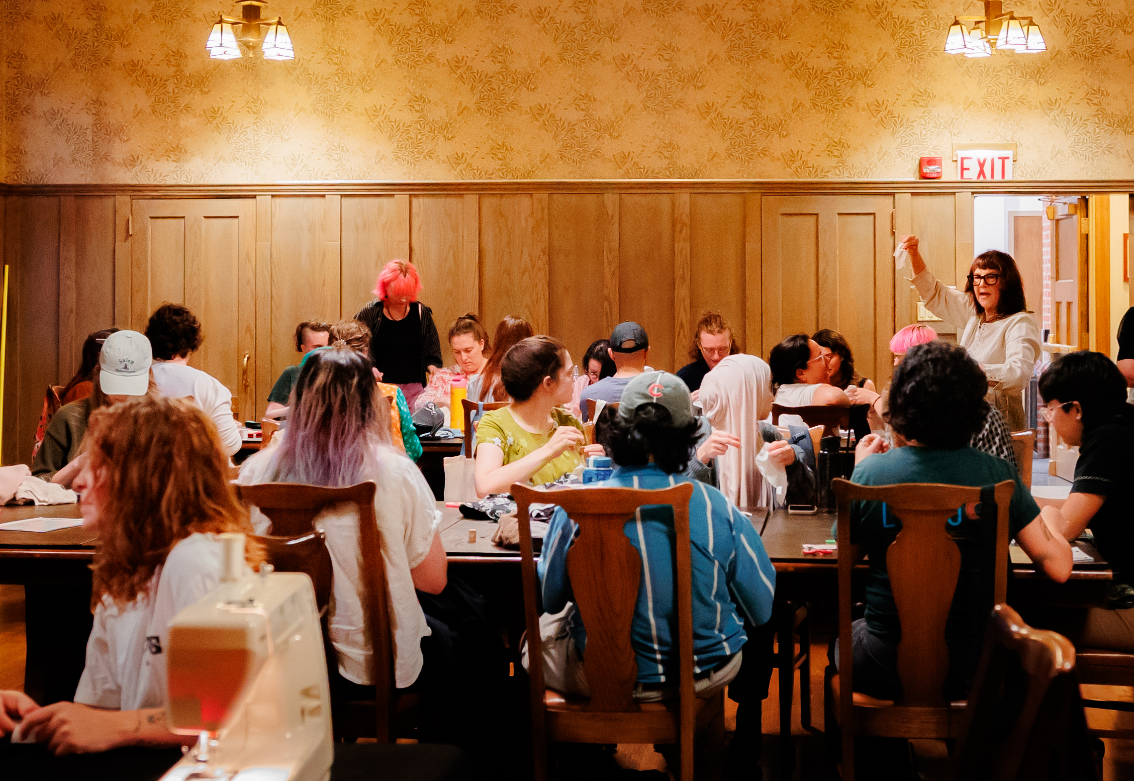 A group of people sits around a long table in a warmly lit room, engaged in a workshop or class, while an instructor stands and gestures at the front. Sewing machines and various materials are visible on the table.