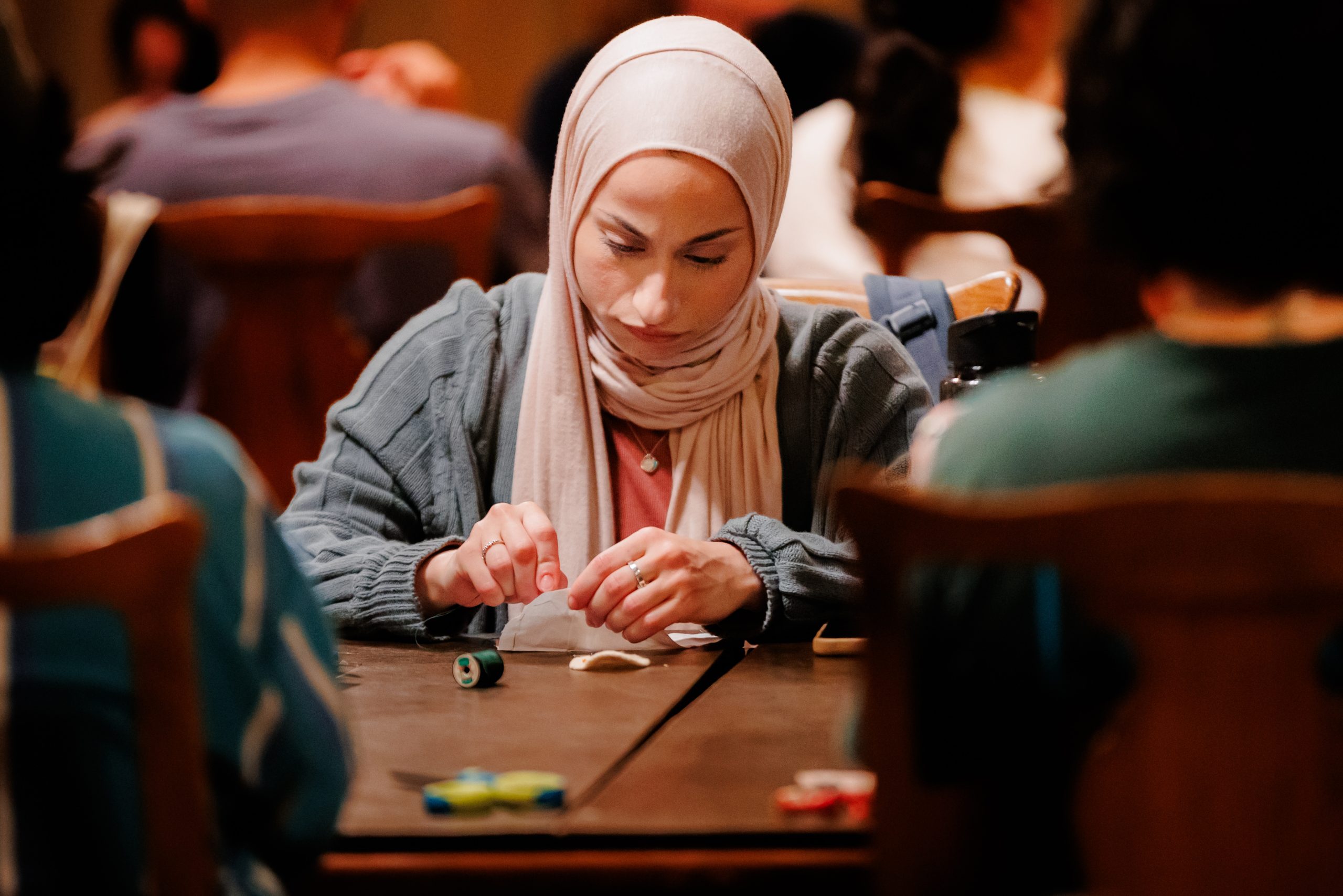 A woman wearing a light-colored hijab and a blue sweater sits at a table, focusing on a craft project with sewing thread and fabric, while other people are seated nearby in a busy indoor setting.