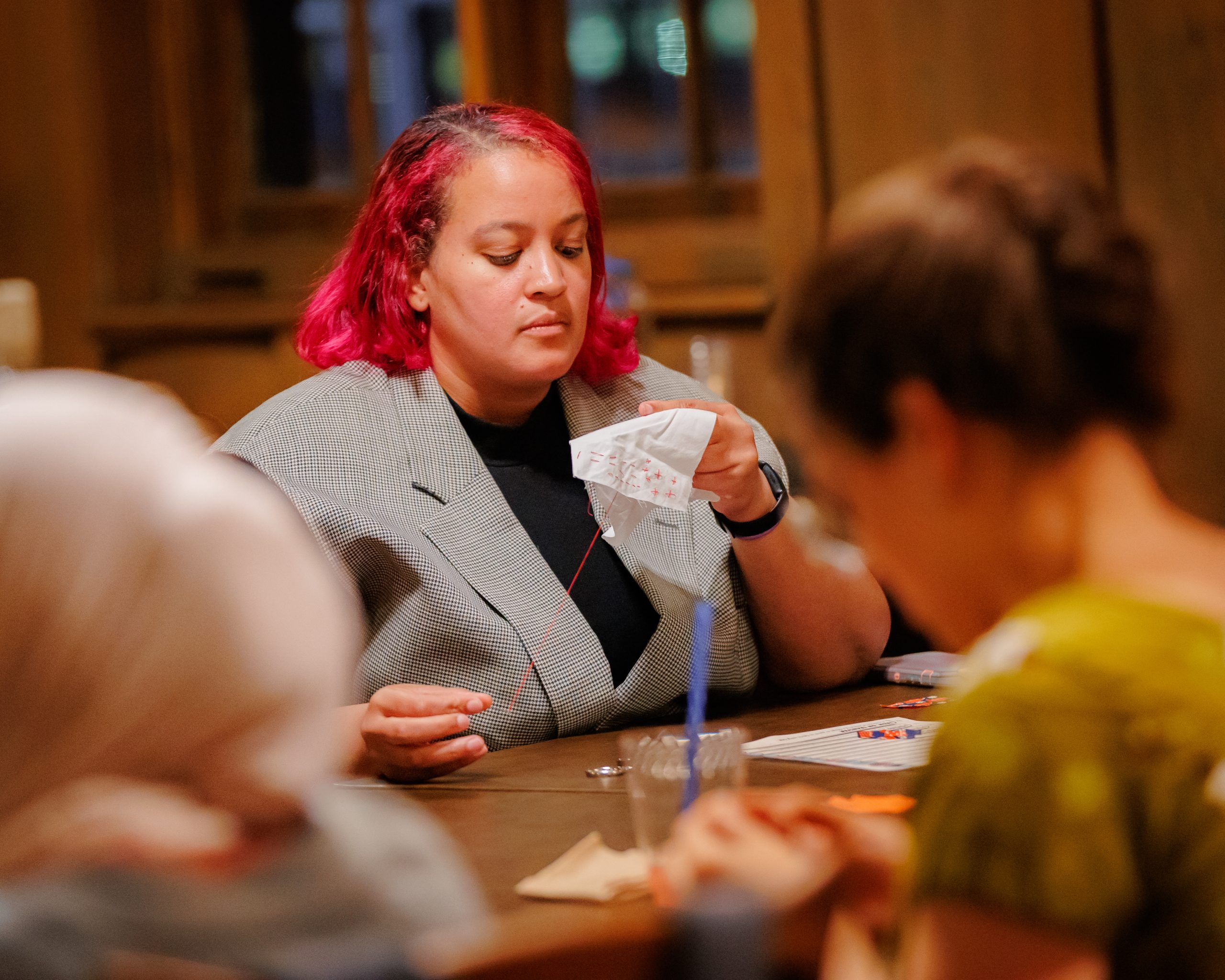 A person with bright pink hair and a gray blazer looks at several pieces of paper while sitting at a table with others in a warmly lit room.