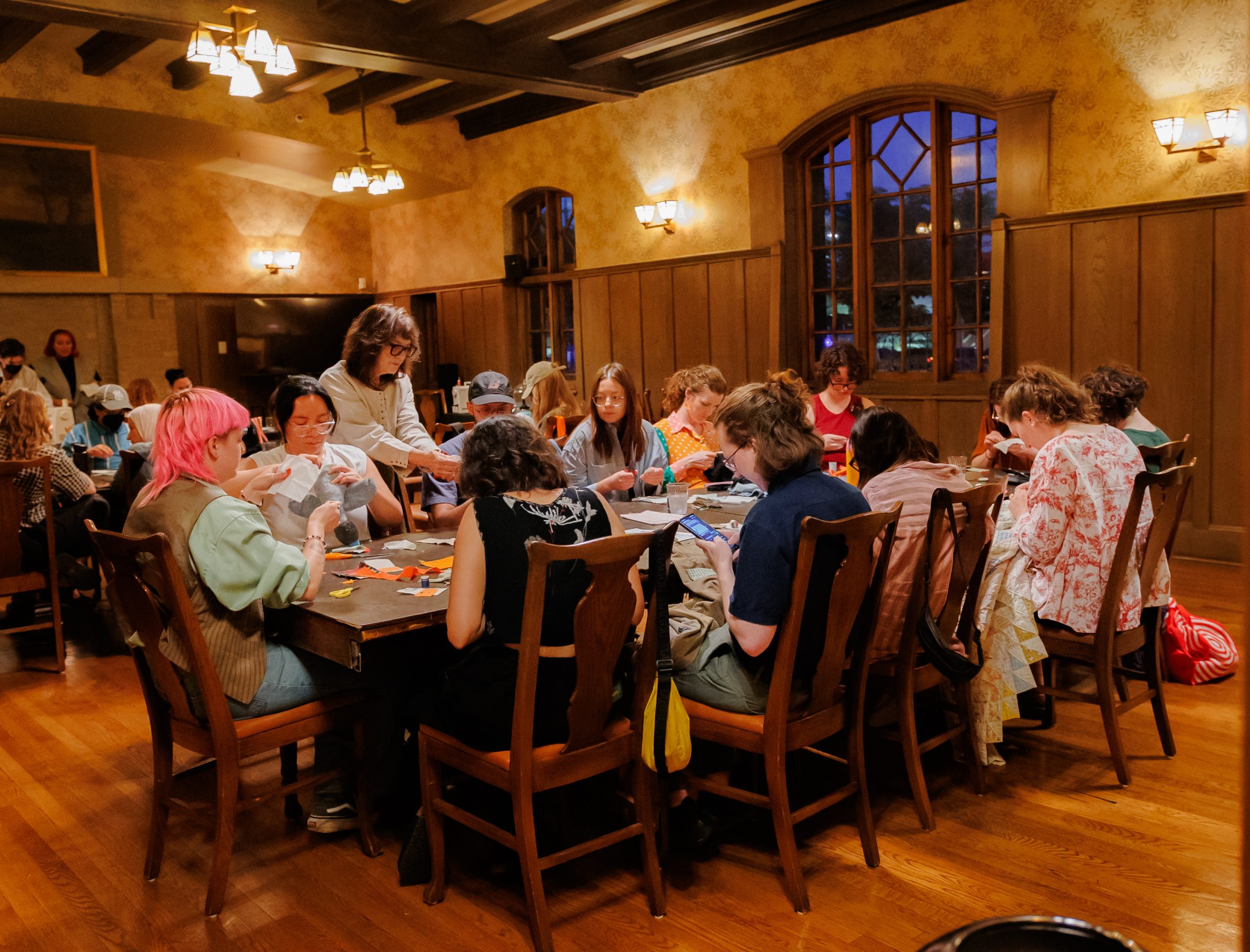 A group of people sit around a large wooden table in a warmly lit room, engaged in crafting activities with fabric and supplies, while a few others stand nearby assisting.