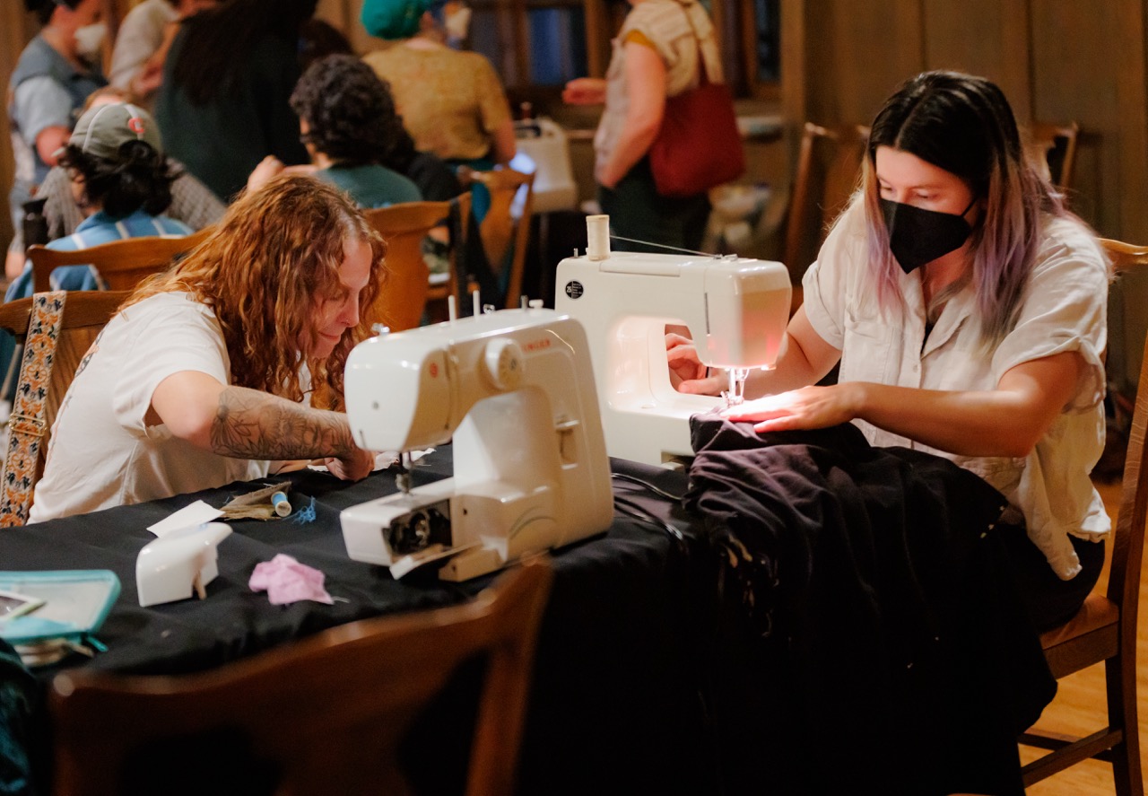 Two people sit at a table using sewing machines, focused on their work. One person wears a black face mask. Several other people are visible in the background, suggesting a group workshop or community event.