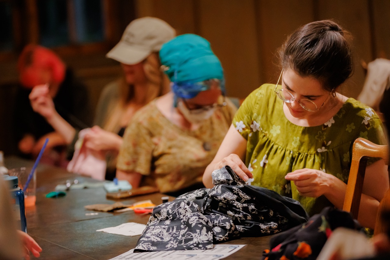 A group of women sit around a wooden table, focused on sewing projects. One woman in the foreground wears glasses and a green dress, stitching black fabric. Scissors, cloth, and sewing supplies are scattered on the table.