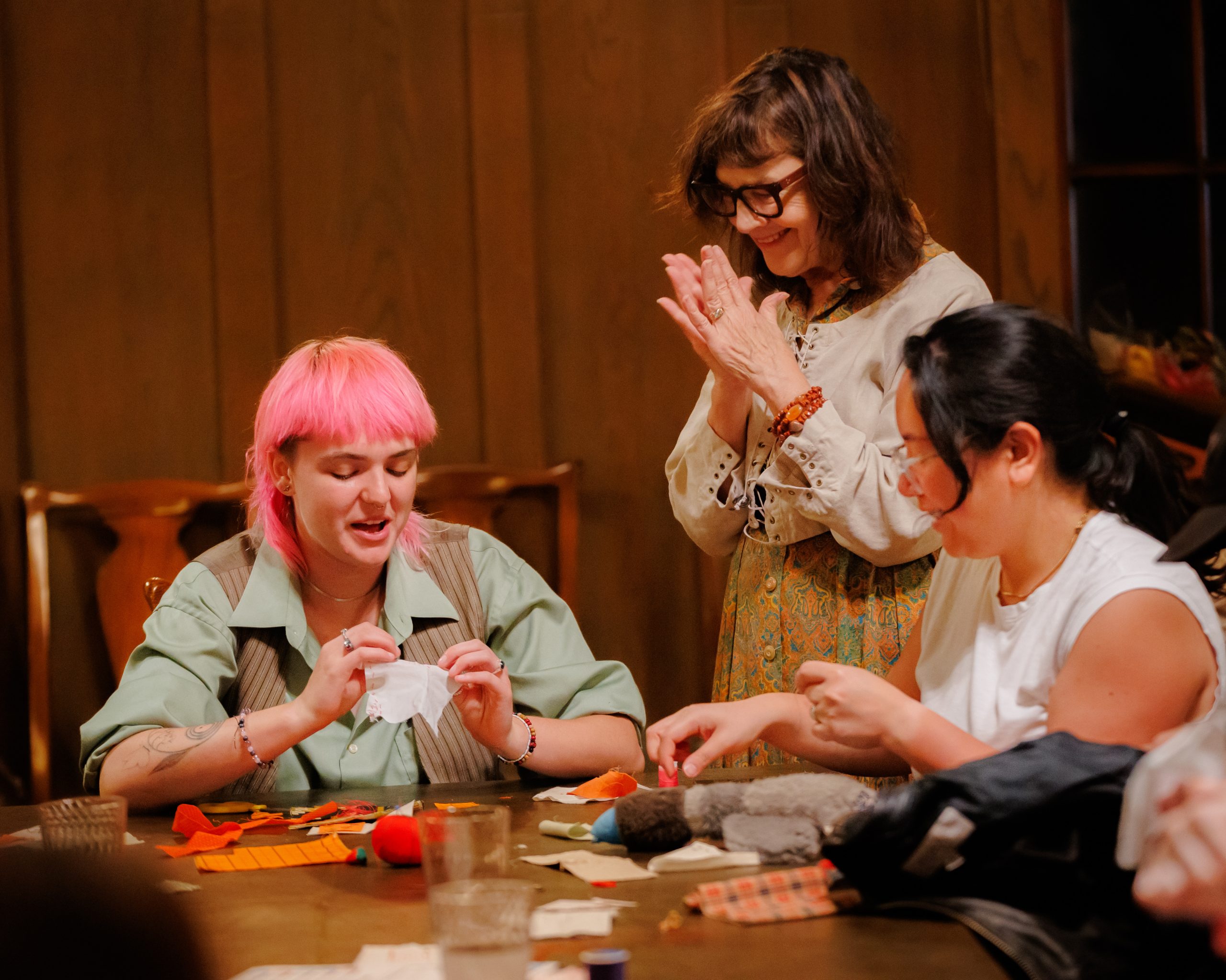 Three women are sitting at a table working on a craft project with fabric pieces. One woman with pink hair is holding a piece, another is sewing, and a third woman stands, smiling and watching them. The setting appears cozy and warm.