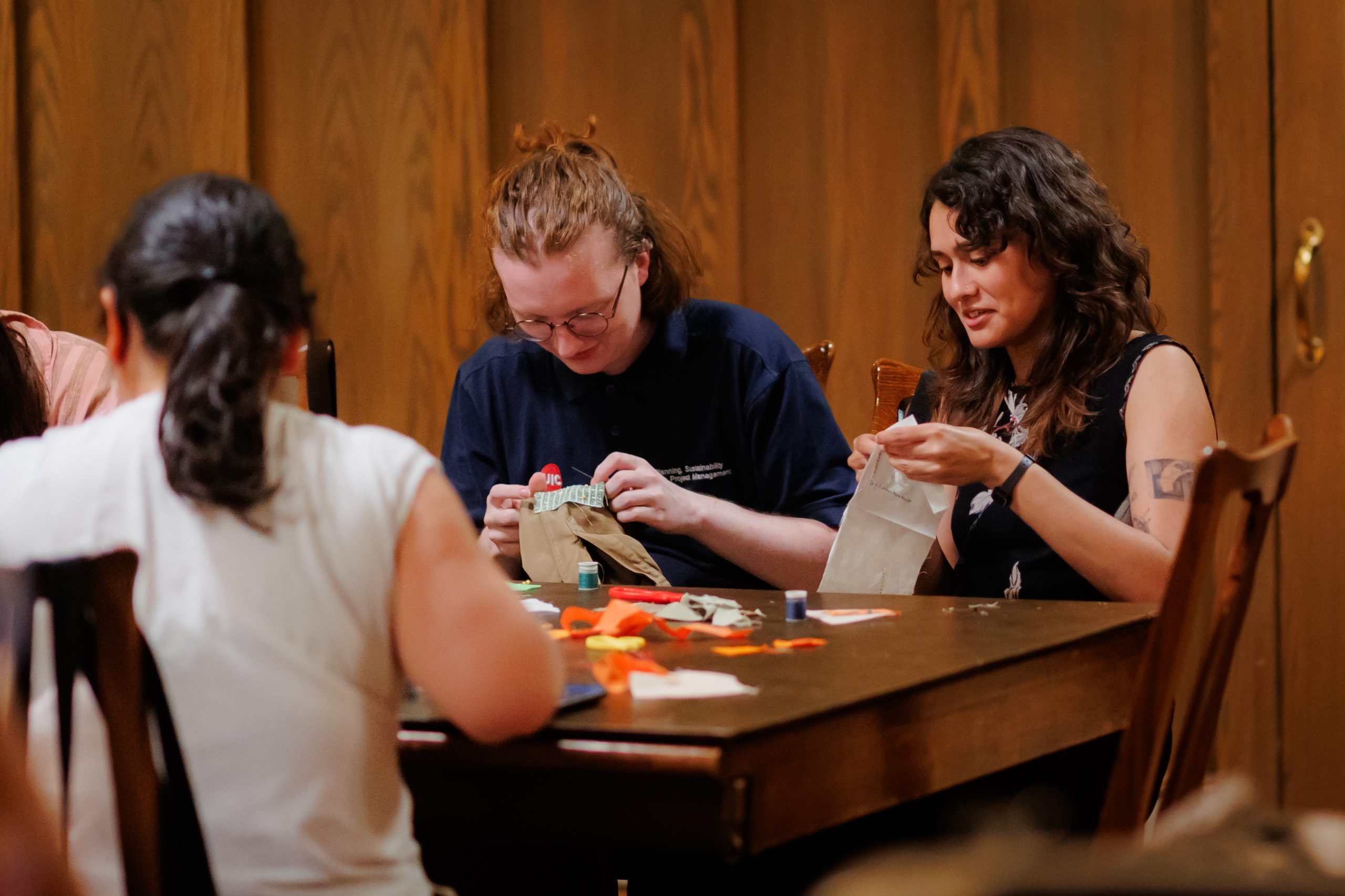 Three people sit around a wooden table working on crafts. Two people are focused on sewing fabric, with various materials and thread on the table. The background is a wood-paneled wall.