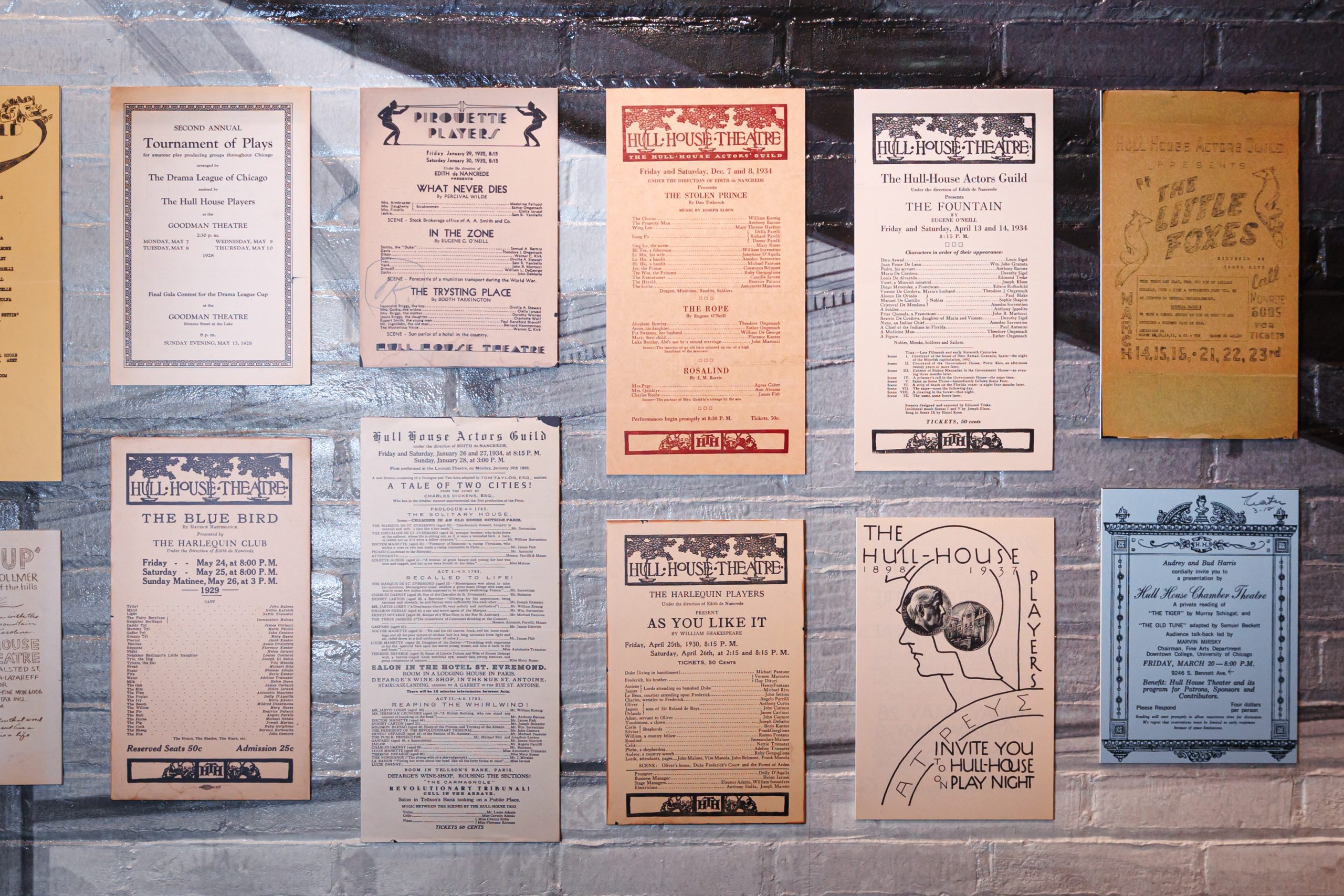 A brick wall displays several vintage theater playbills and posters in various colors and fonts, advertising past performances and events by the Hull House Players and other theater groups.