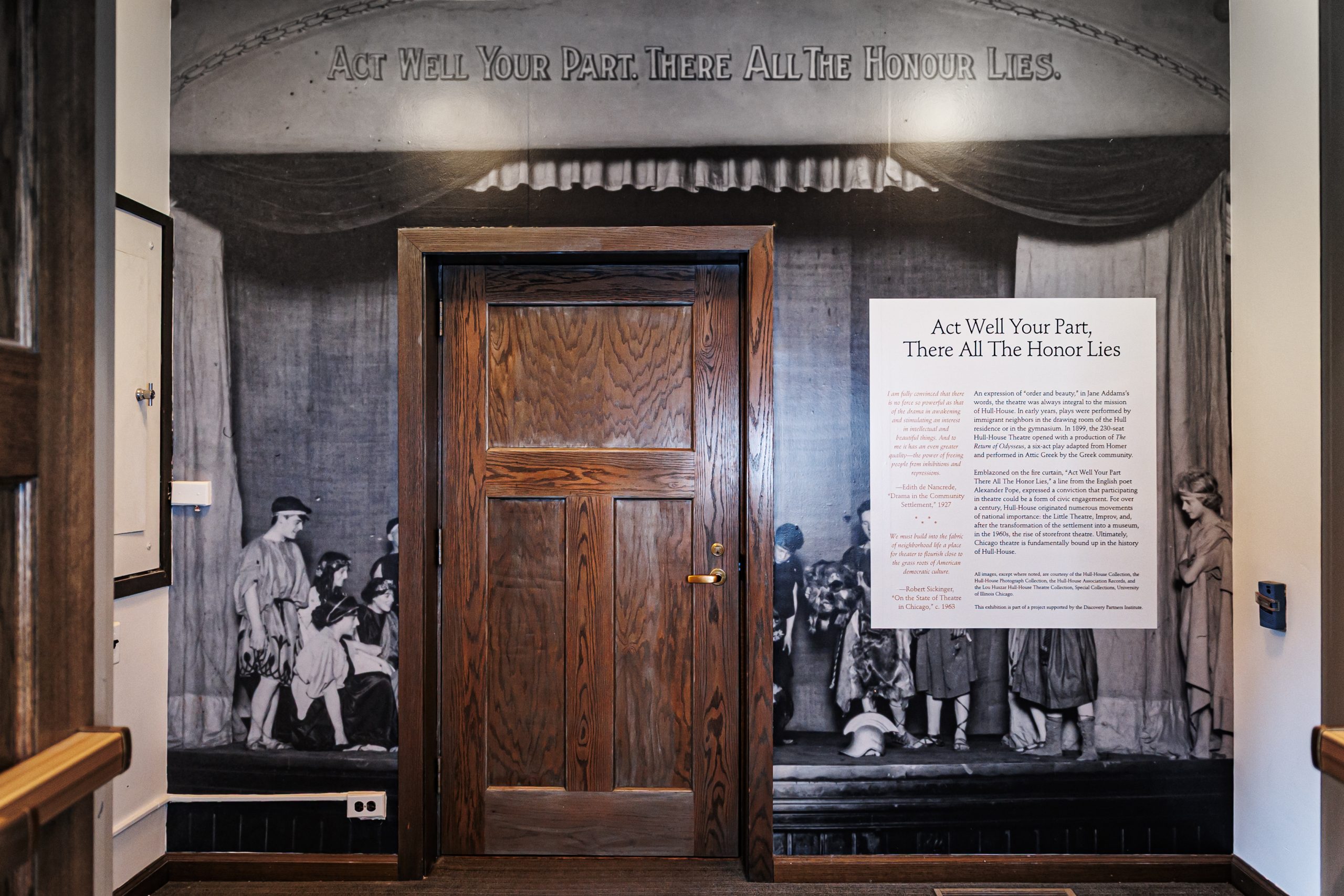 A wooden door is set into a wall covered with a historic black-and-white photo of children on a stage. Above the door is the phrase Act Well Your Part There All The Honour Lies and a display panel with text to the right.