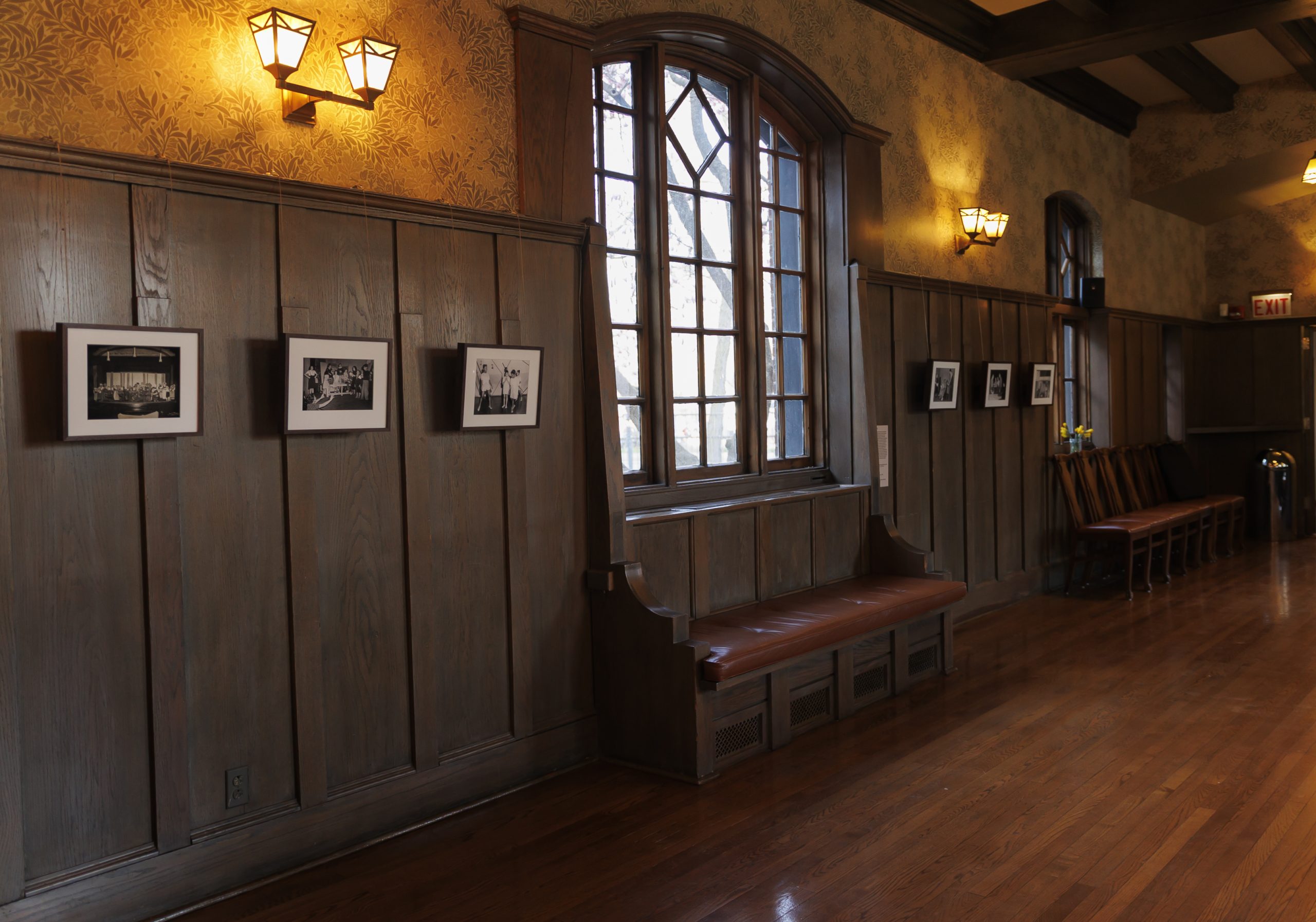 A hallway with wood-paneled walls, framed black-and-white photos, a large arched window with a wooden bench beneath it, wall lamps, and a row of chairs along the polished wooden floor.