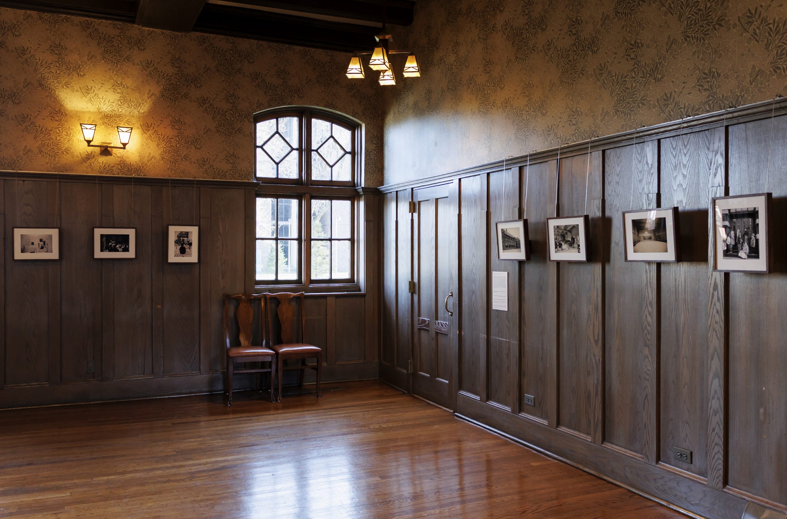 A quiet, empty room with wooden floors, wood-paneled walls, a large window, two wooden chairs, and black-and-white photos framed and hung on the walls under warm lighting.