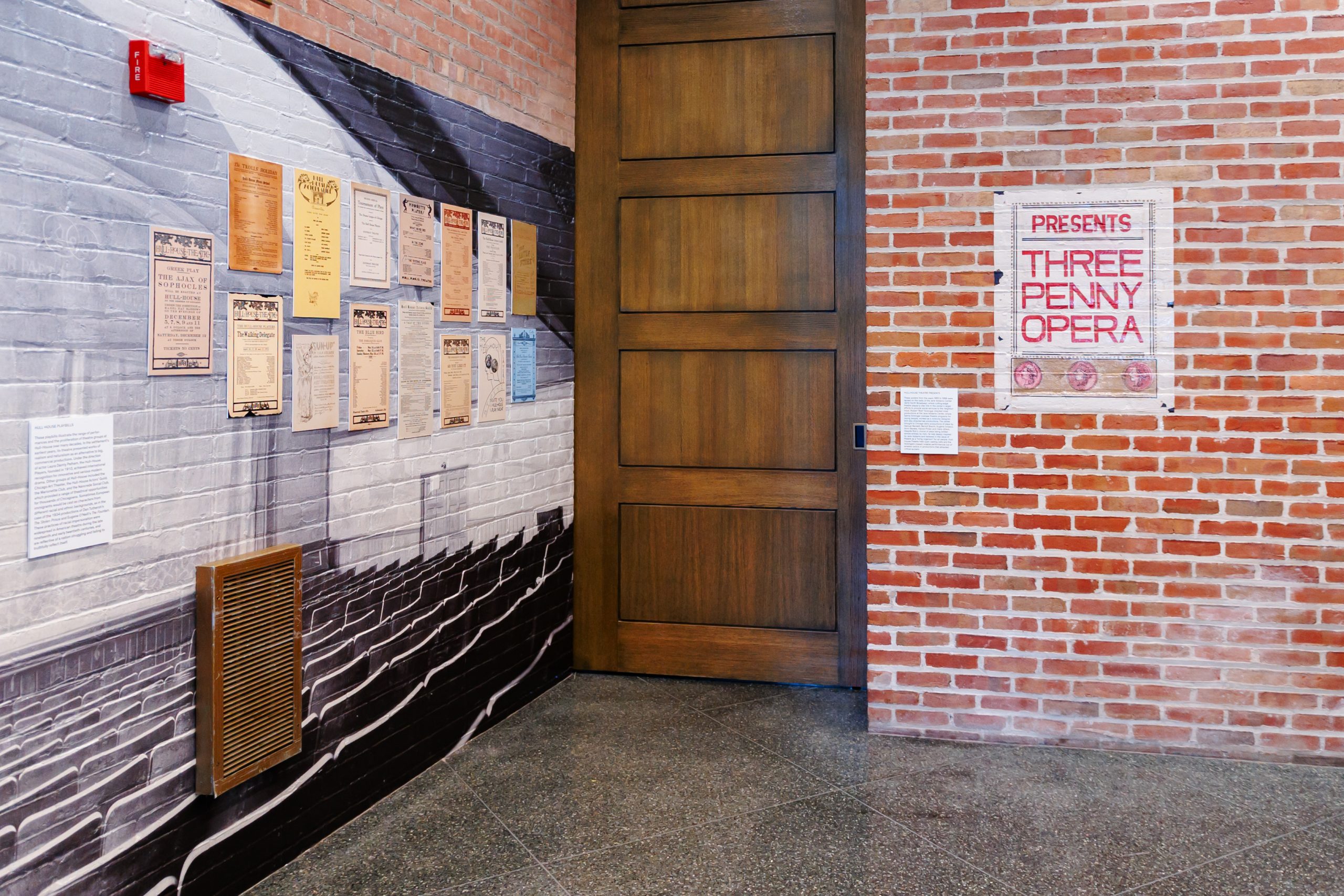 A brick wall with posters and playbills on the left, a large wooden door in the center, and a framed sign on the right that reads Presents Three Penny Opera. The floor is polished stone.